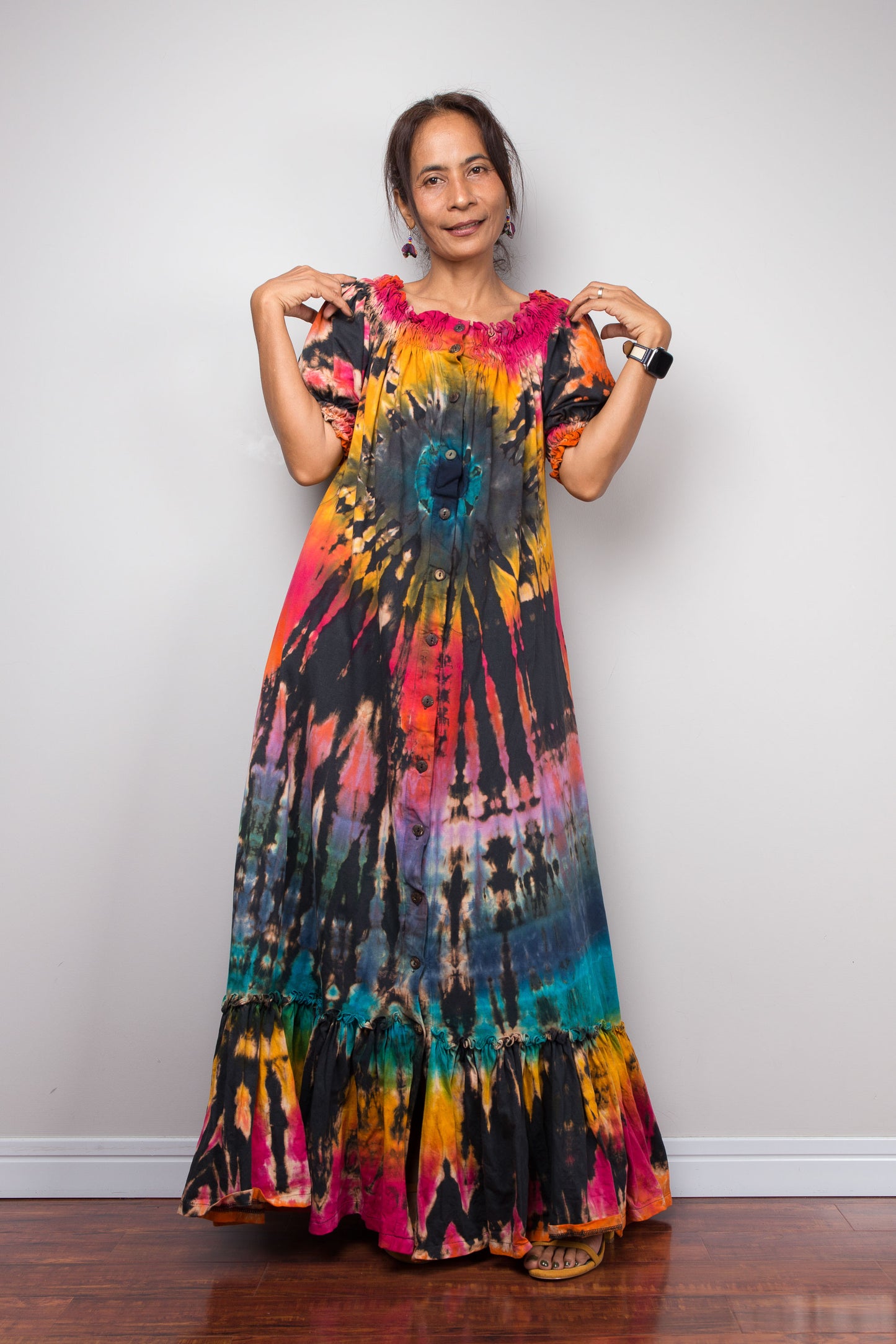 A woman modeling a short sleeved Nuichan maxi dress with a vibrant tie-dye pattern in shades of orange, yellow, pink, purple, blue, and black, standing against a plain light gray wall.