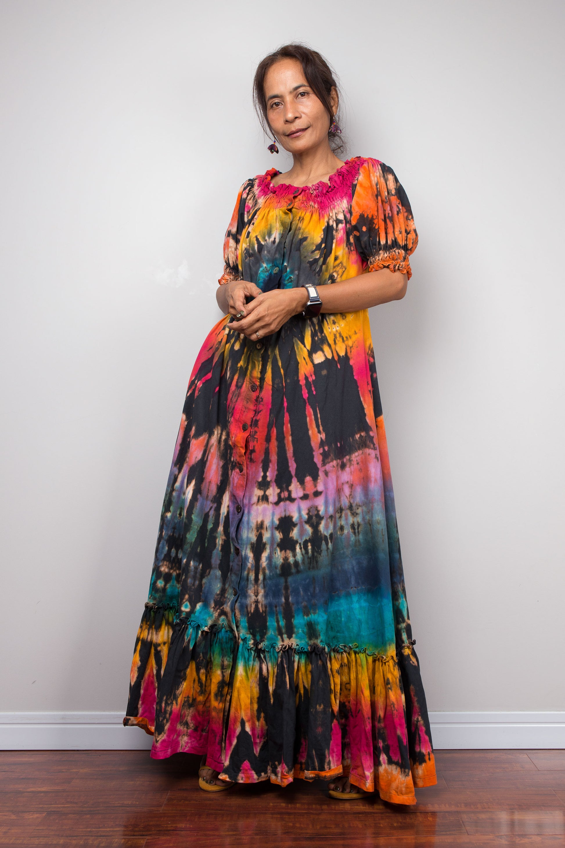 A petite woman wearing a vibrant, multicolor Nuichan tie dye maxi shirt dress with short puff sleeves and a tiered ruffled hem, standing against a plain light gray wall on a wooden floor.