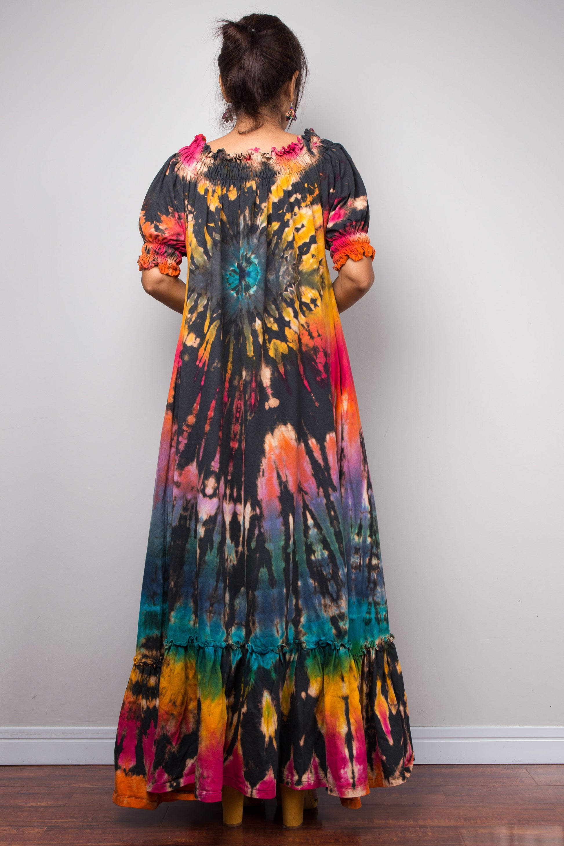 A woman viewed from behind wearing a long-sleeved maxi dress with a vibrant tie-dye pattern in black, orange, yellow, pink, and teal, standing against a plain light gray wall.
