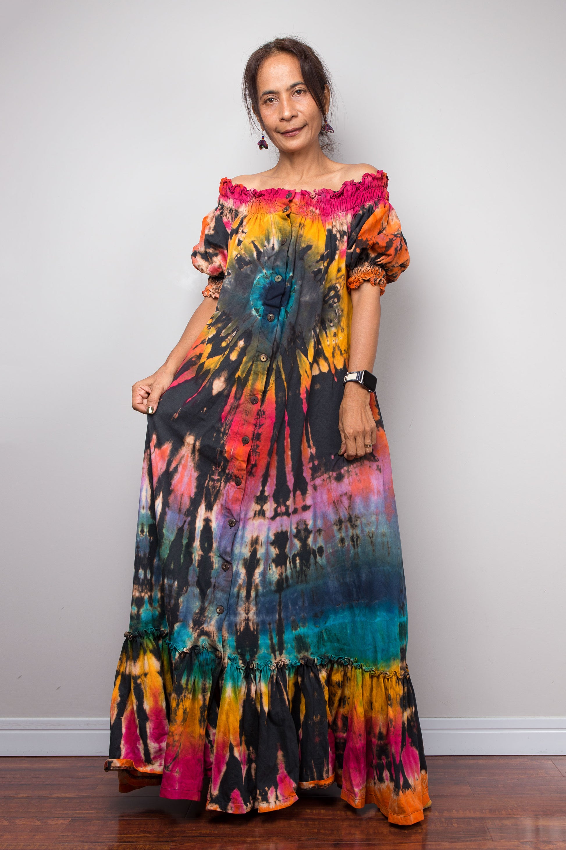 A petite woman modeling a vibrant, multicolor tie dye shirt maxi dress with puff sleeves and an off-the-shoulder neckline, standing against a plain light gray wall.