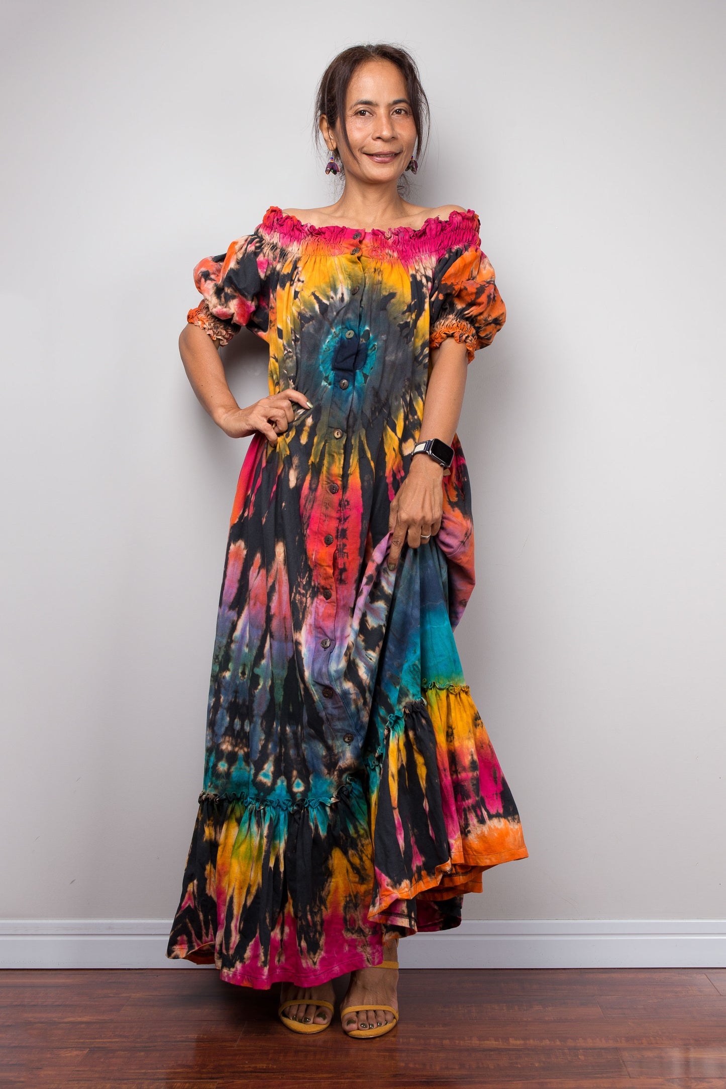 A woman modeling a vibrant tie-dye maxi dress with puff sleeves, standing against a plain light gray wall on a dark wood floor.