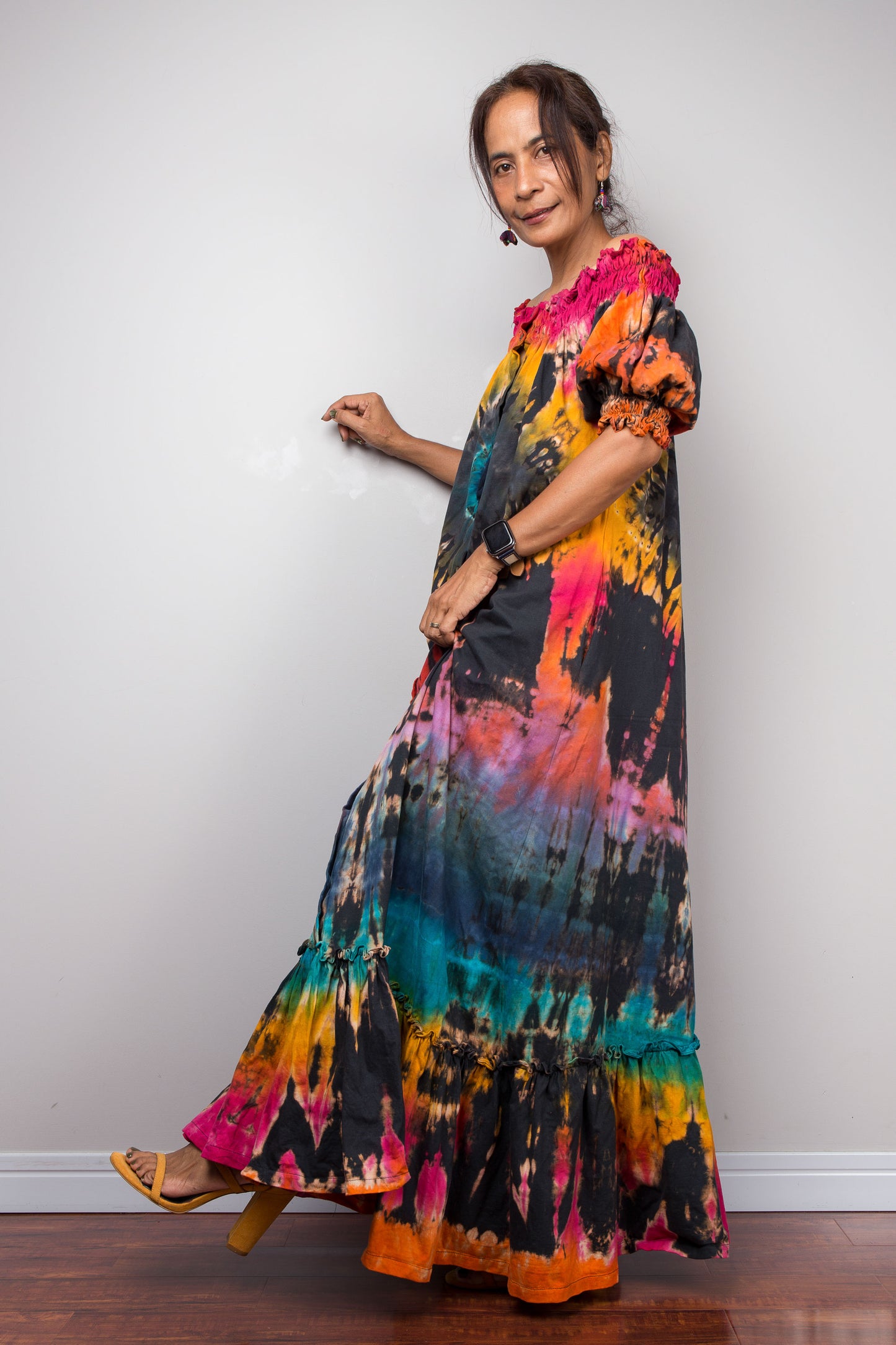 A petite woman wearing a long, colorful Nuichan tie dye maxi shirt dress with puff sleeves and a tiered ruffled hem, standing against a plain light gray wall.
