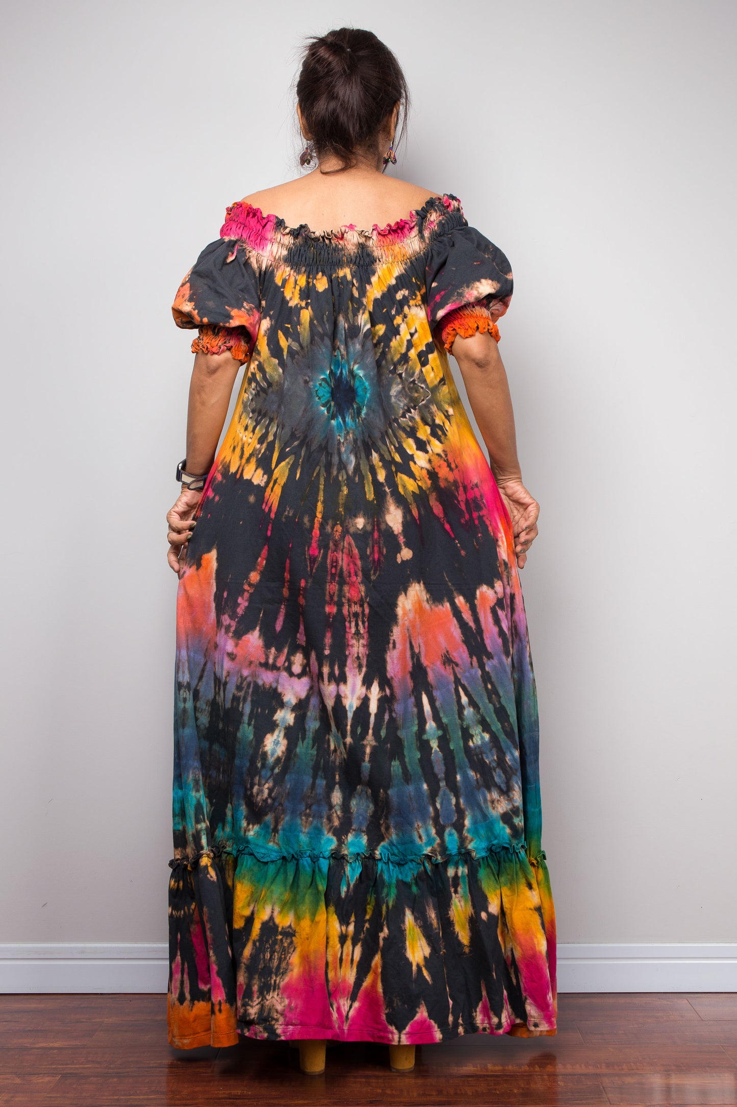 A petite woman viewed from behind wearing a long off-the-shoulder maxi dress with a vibrant tie-dye pattern in black, orange, pink, yellow, and teal, standing against a plain light gray wall.