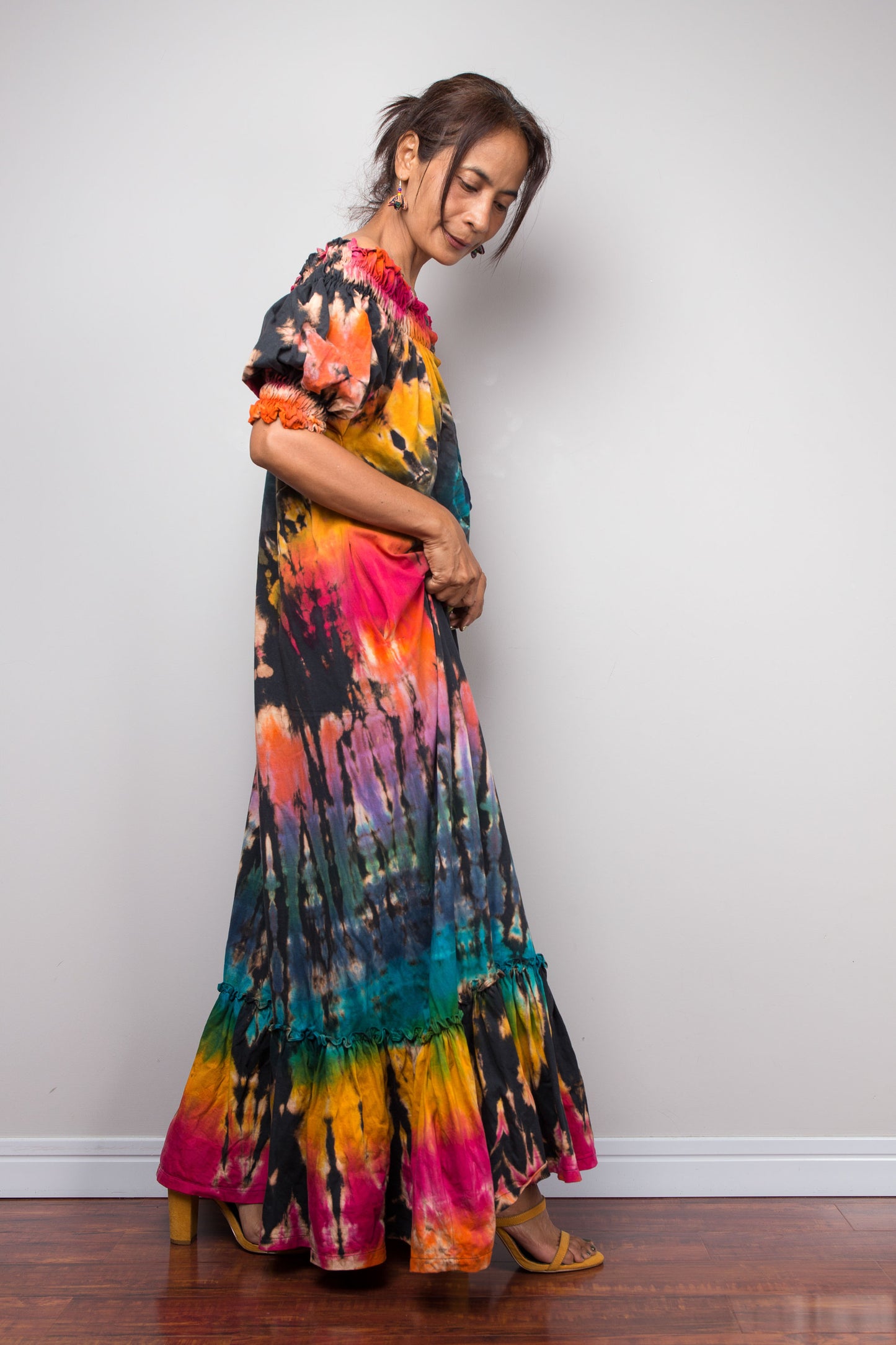 A woman wearing a long-sleeved maxi dress with a vibrant tie-dye pattern in pink, orange, yellow, teal, and black, standing against a plain light gray wall.