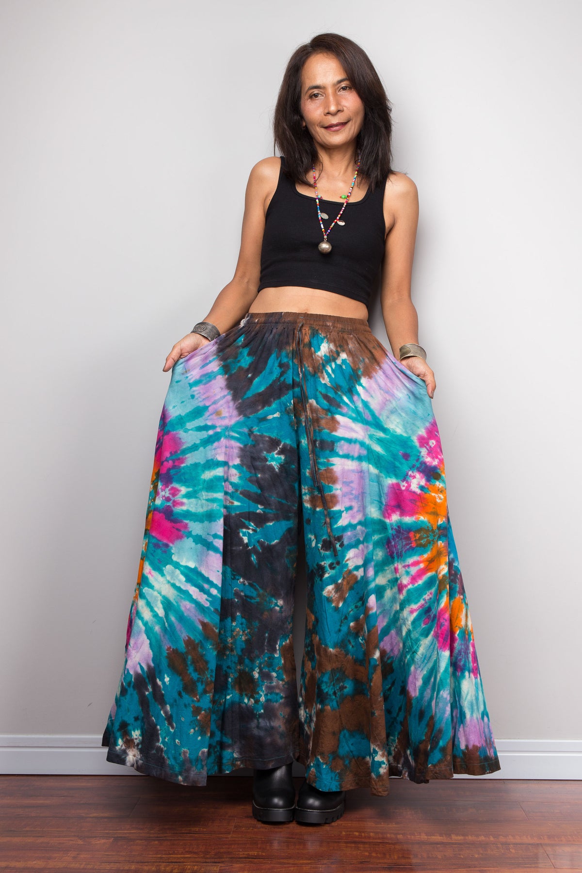 Woman holding the teal tie dye palazzo pants open at arm's length, showing the dramatic wide-leg spread and skirt-like silhouette of the boho trousers