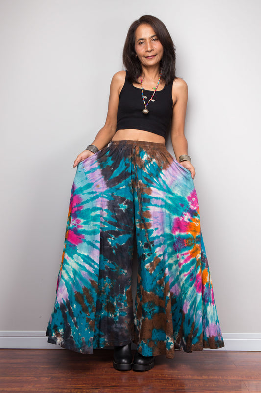 Woman holding the teal tie dye palazzo pants open at arm's length, showing the dramatic wide-leg spread and skirt-like silhouette of the boho trousers