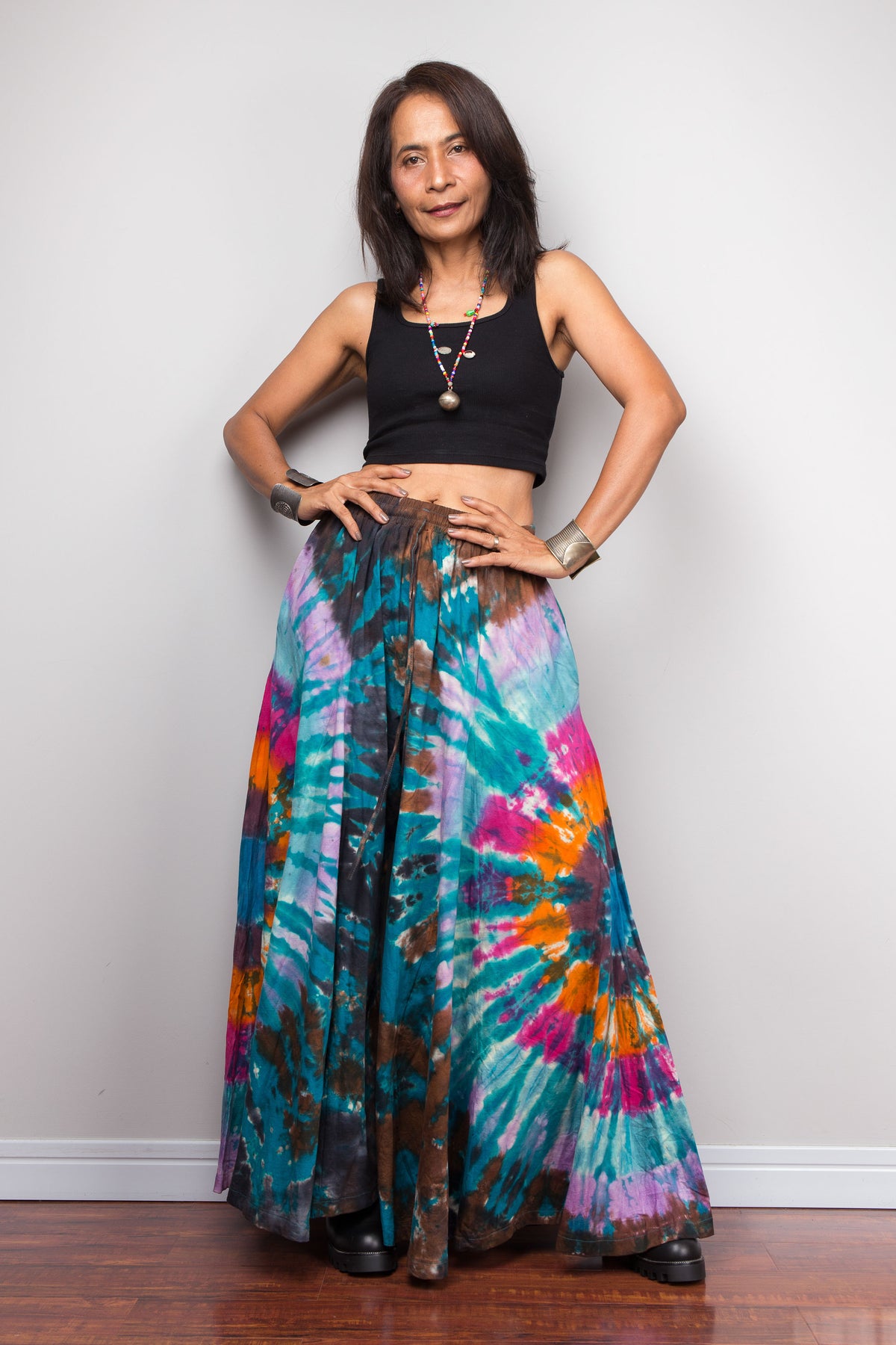 Woman standing with hands on hips wearing Nuichan teal spiral tie dye wide leg palazzo pants, front view showing the full mandala pattern radiating from the waist to the hem