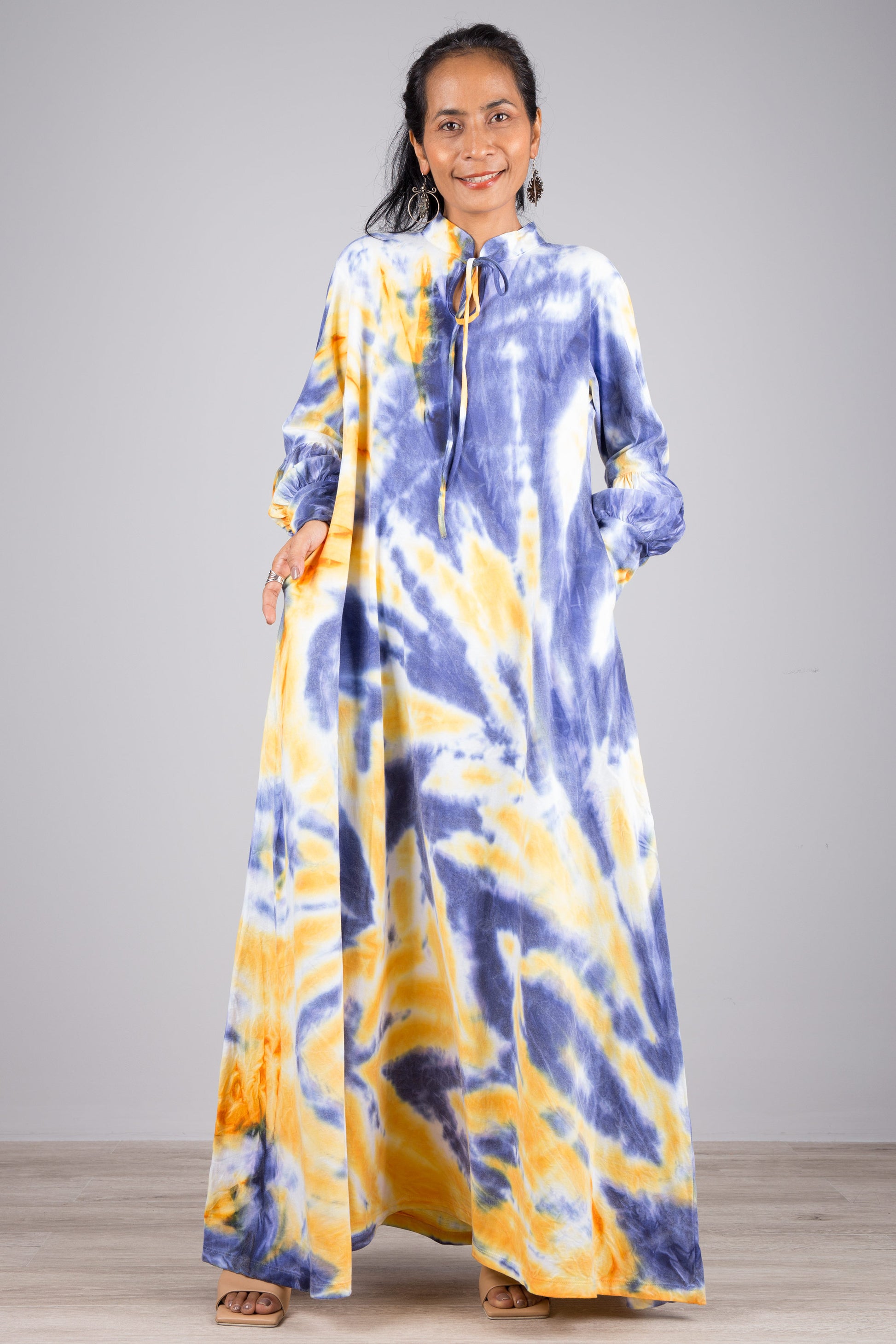 Blue and yellow tie-dye kaftan maxi dress on a woman, featuring a flowing silhouette, high neck with drawstring, and long sleeves, set against a minimalist gray and wood background, with nude sandals.

