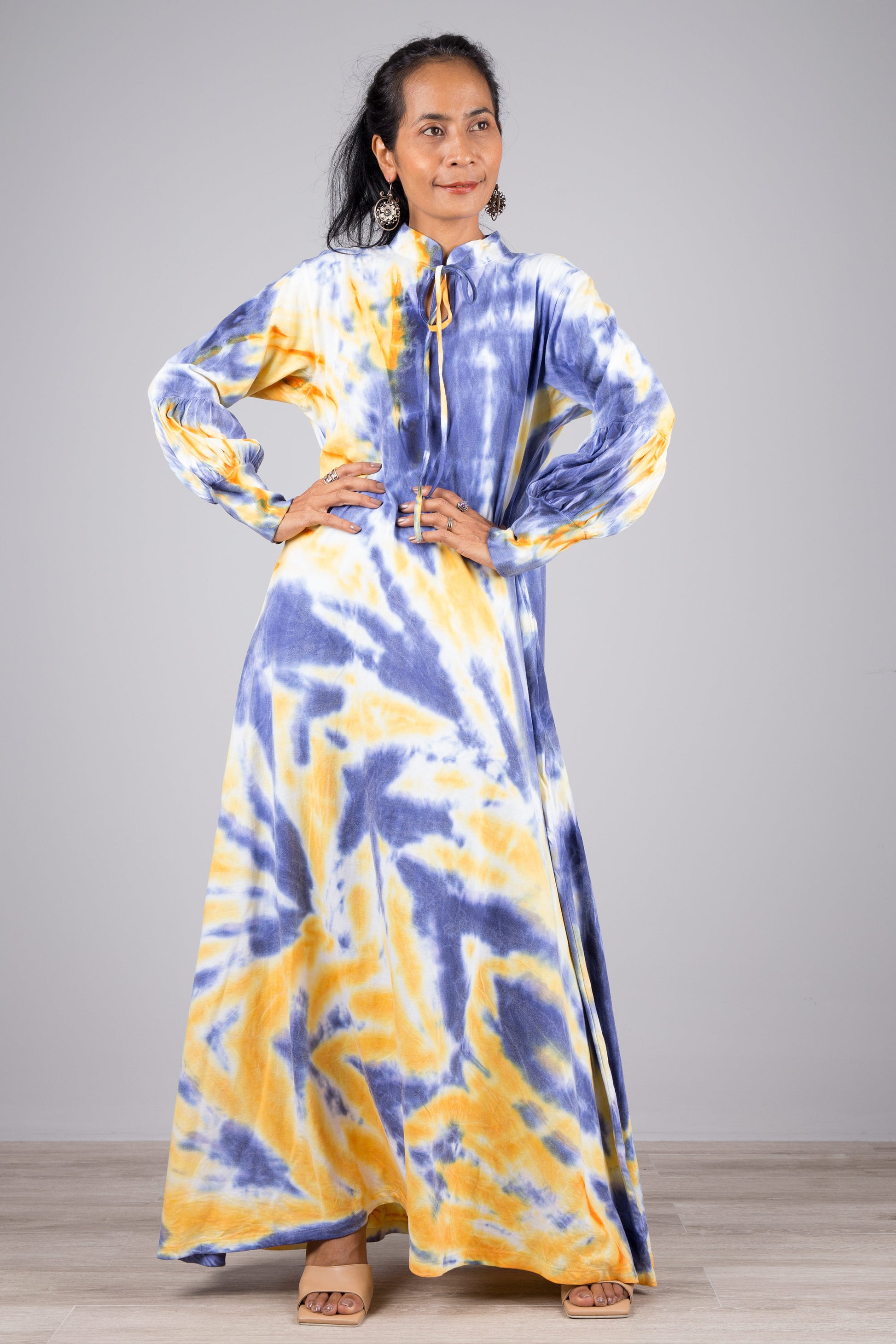 A woman wearing a blue and yellow tie-dye maxi dress with a high neckline and long sleeves, standing against a gray wall with a wooden floor, accessorized with nude sandals and dangling earrings.

