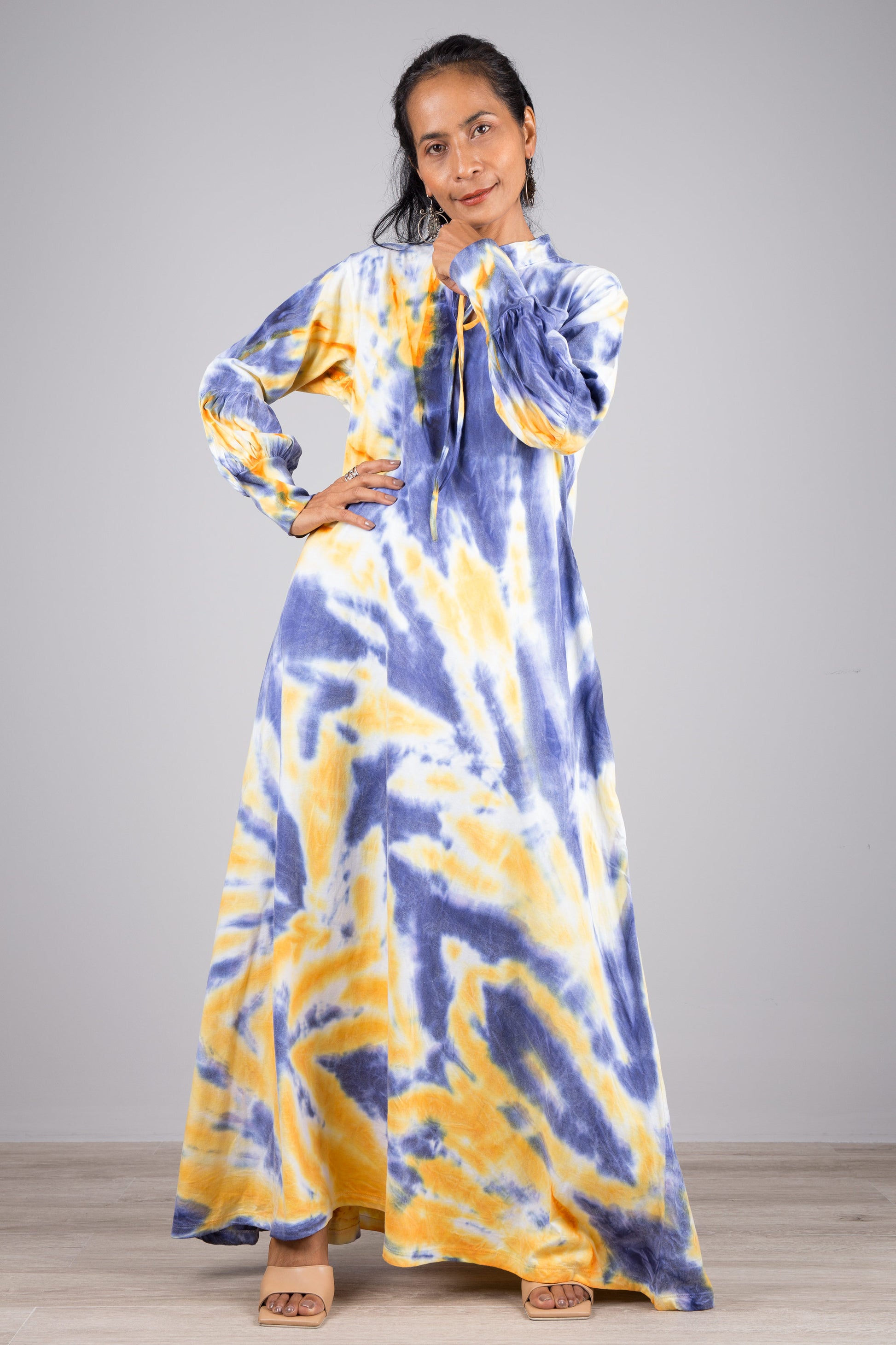 Stylish woman in a floor-length blue and yellow tie-dye kaftan dress with a high neckline and billowy sleeves, posed against a gray wall and wooden floor, wearing nude sandals and earrings.

