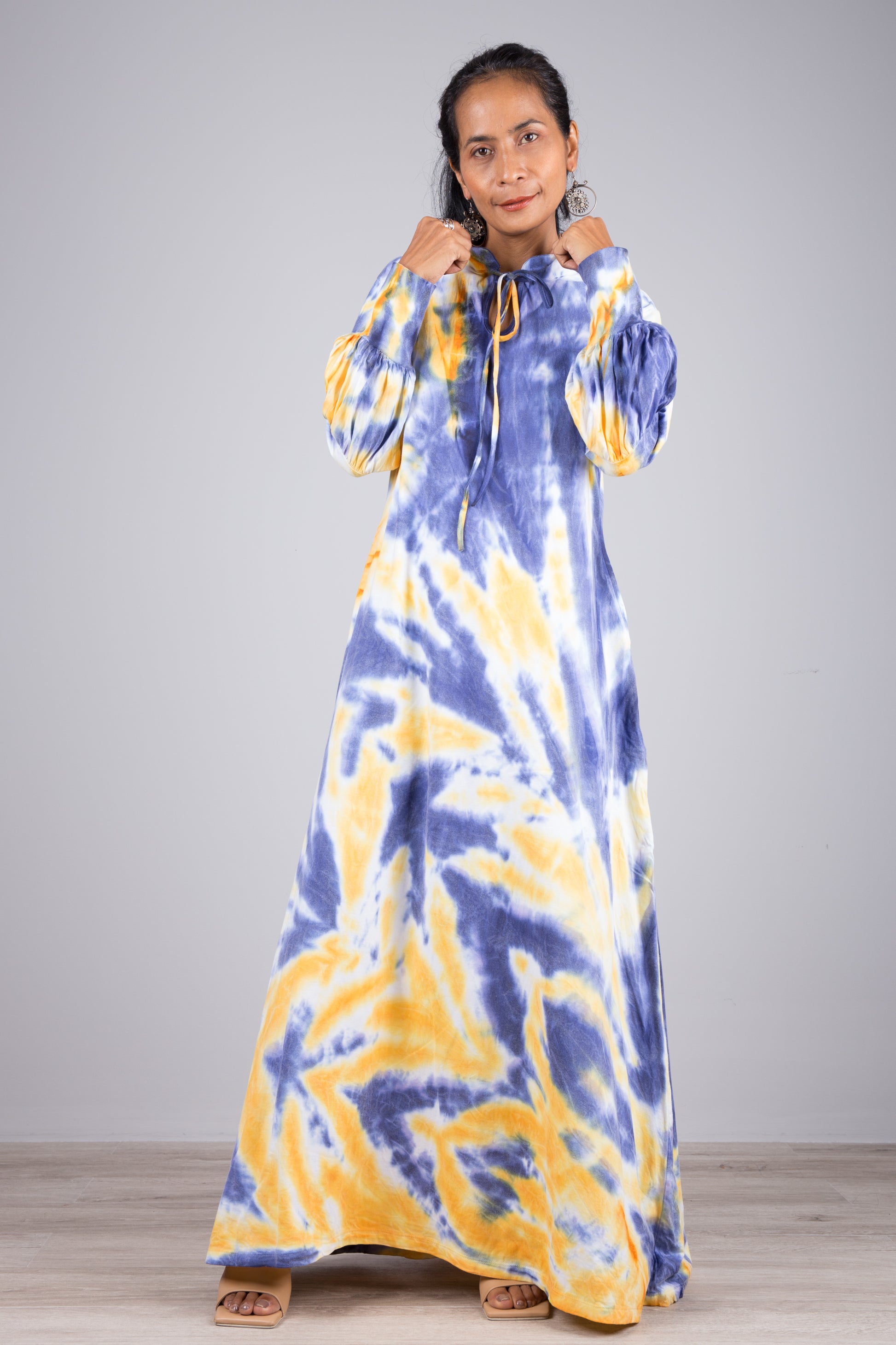 Nuichan blue and yellow tie-dye maxi dress with a high neckline, drawstring tie, and long sleeves, modeled by a woman with nude sandals and dangling earrings, against a simple gray and wood backdrop.

