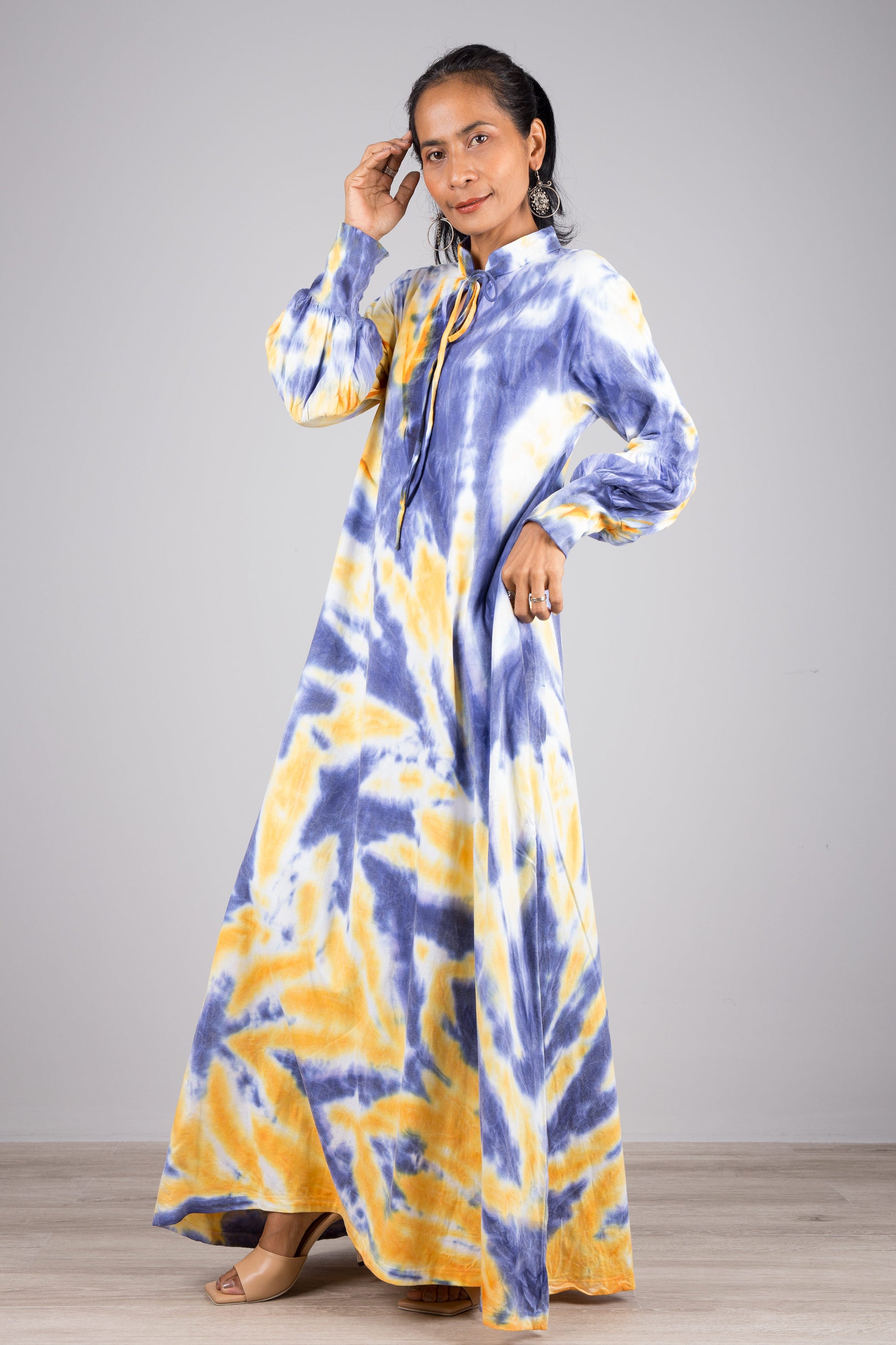 Woman showcasing a blue and yellow tie-dye kaftan maxi dress with a flowing design, high neck, and long sleeves, accessorized with nude sandals and a ring, on a gray and wooden floor setting.

