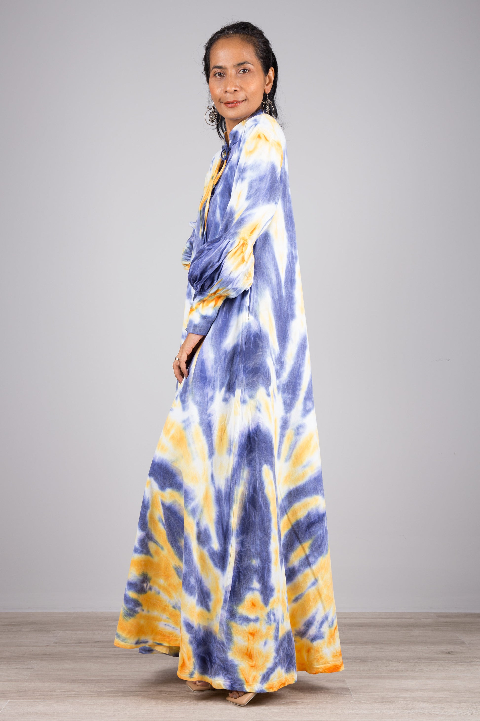 Elegant blue and yellow tie-dye kaftan maxi dress with a high neckline and gathered sleeves, worn by a woman against a gray wall and wooden floor, paired with nude sandals and dangling earrings.

