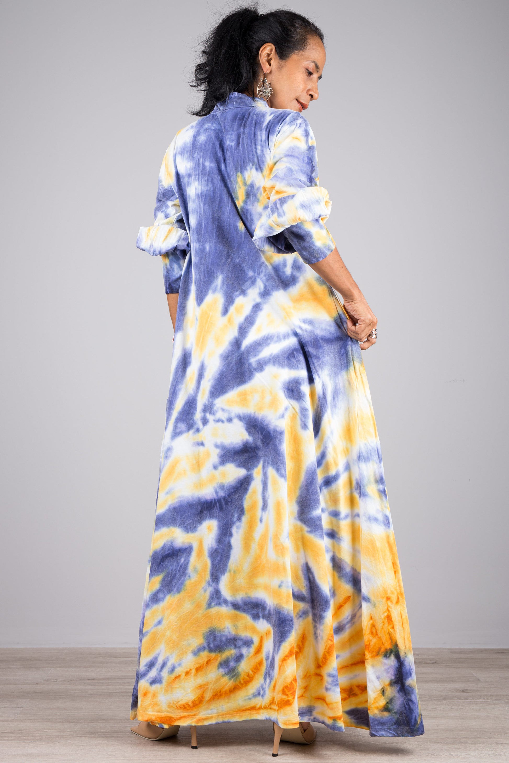 Model in a blue and yellow tie-dye pattern kaftan maxi dress featuring a high neck with drawstring and long sleeves, standing on a wooden floor with a gray wall background, wearing nude sandals."

