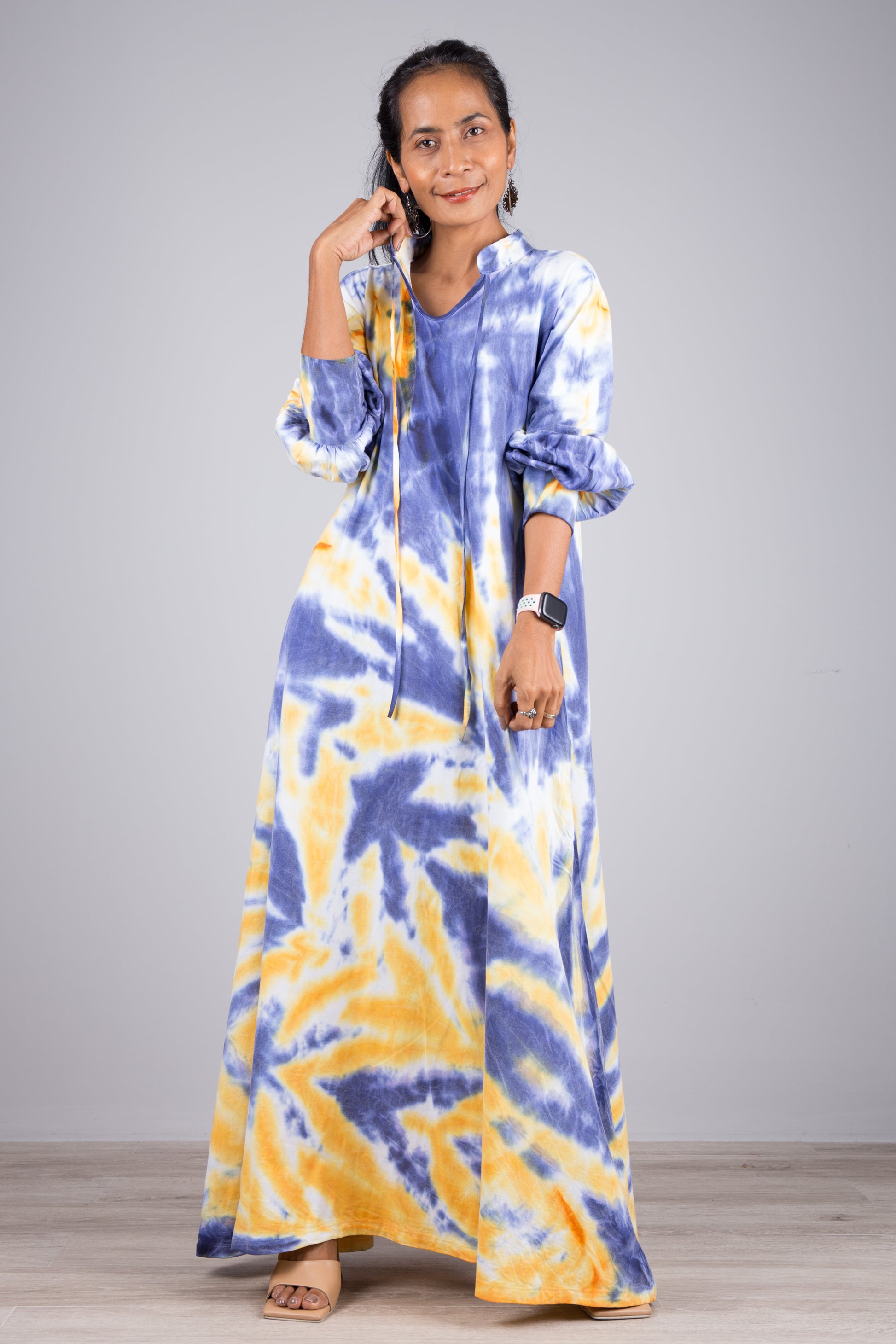 Woman in a vibrant blue and yellow tie-dye kaftan maxi dress with a high neckline and billowy sleeves, posed against a gray wall and wooden floor, styled with nude sandals and simple jewelry.

