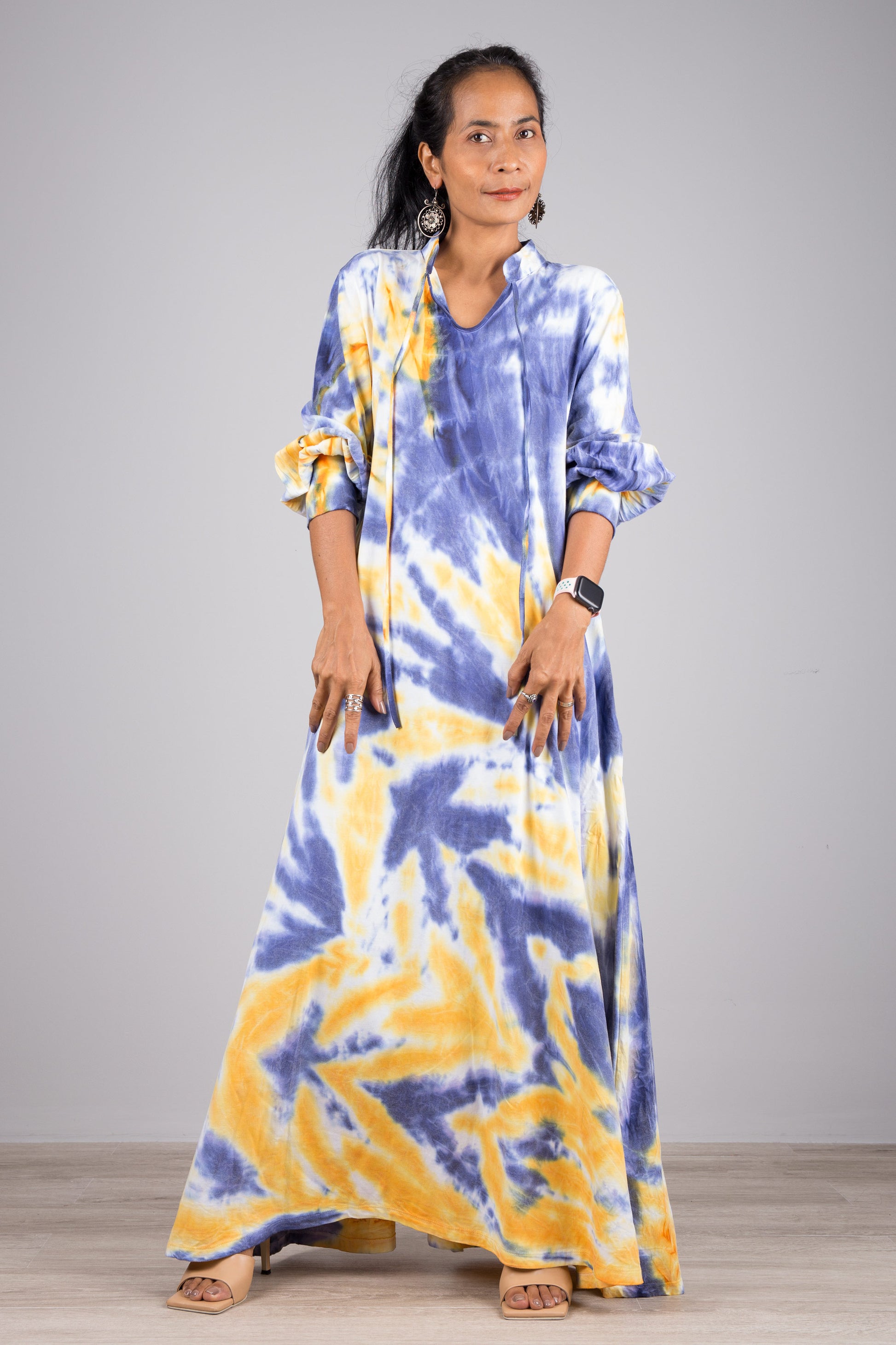 Blue and yellow tie-dye kaftan maxi dress with a high neck and long sleeves, worn by a woman with nude sandals and dangling earrings, set against a minimalist gray and wood interior.

