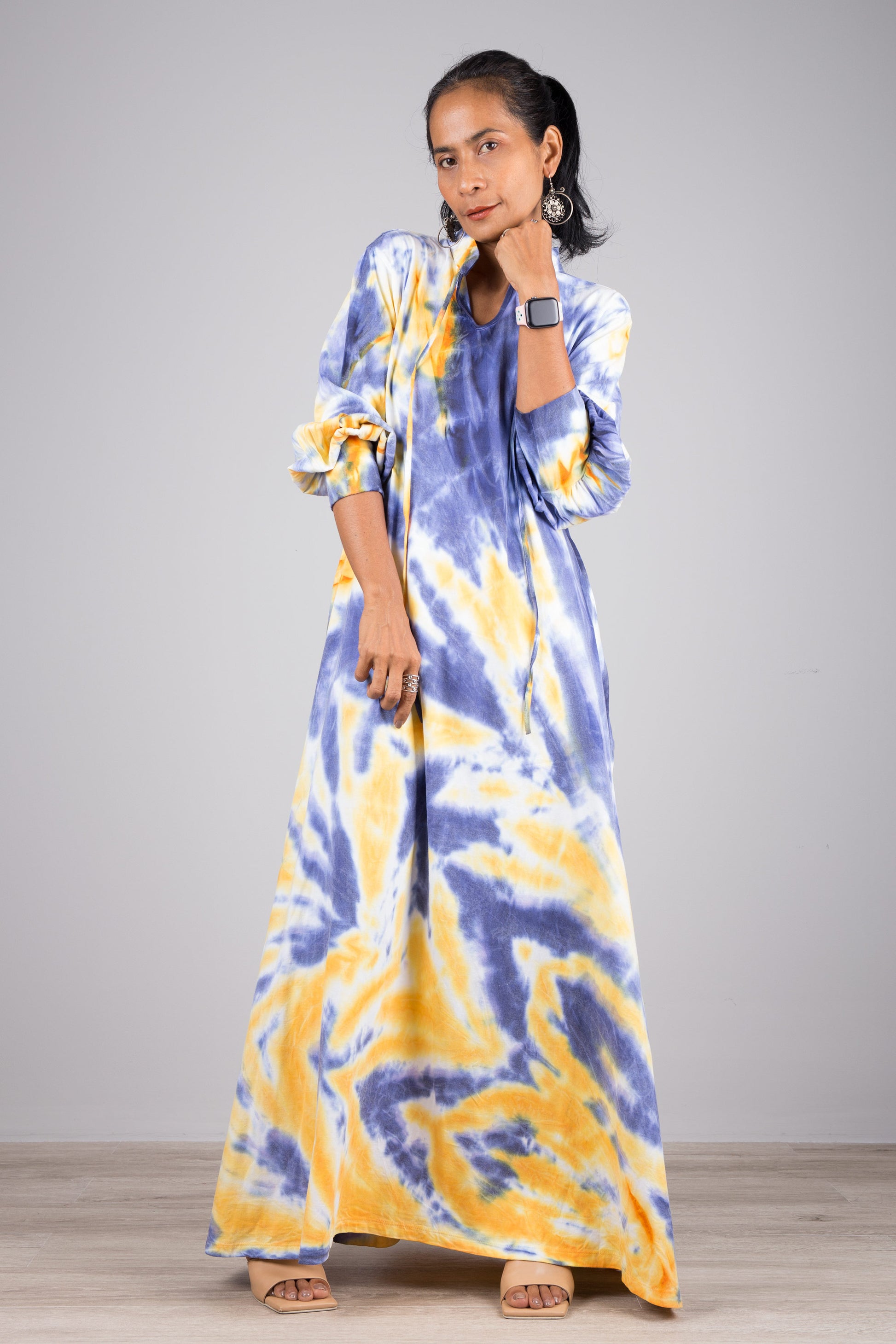 Fashionable woman wearing a blue and yellow tie-dye kaftan maxi dress with a drawstring neckline and long sleeves, standing on a wooden floor with a gray wall backdrop, accessorized with nude sandals.

