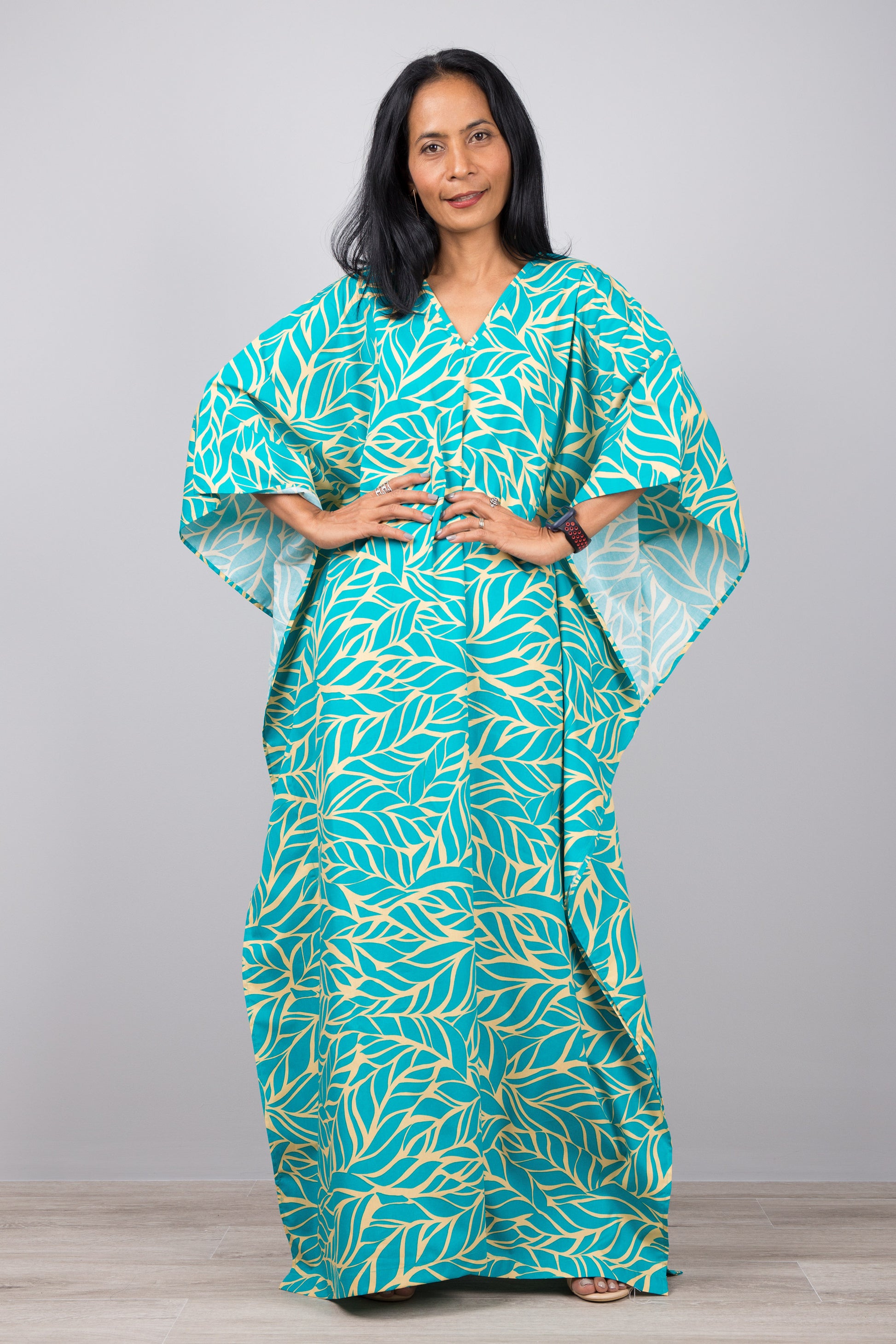 Woman in a turquoise leaf-patterned kaftan dress with yellow accents, standing against a gray wall with a wooden floor, accessorized with a smartwatch and silver ring.

