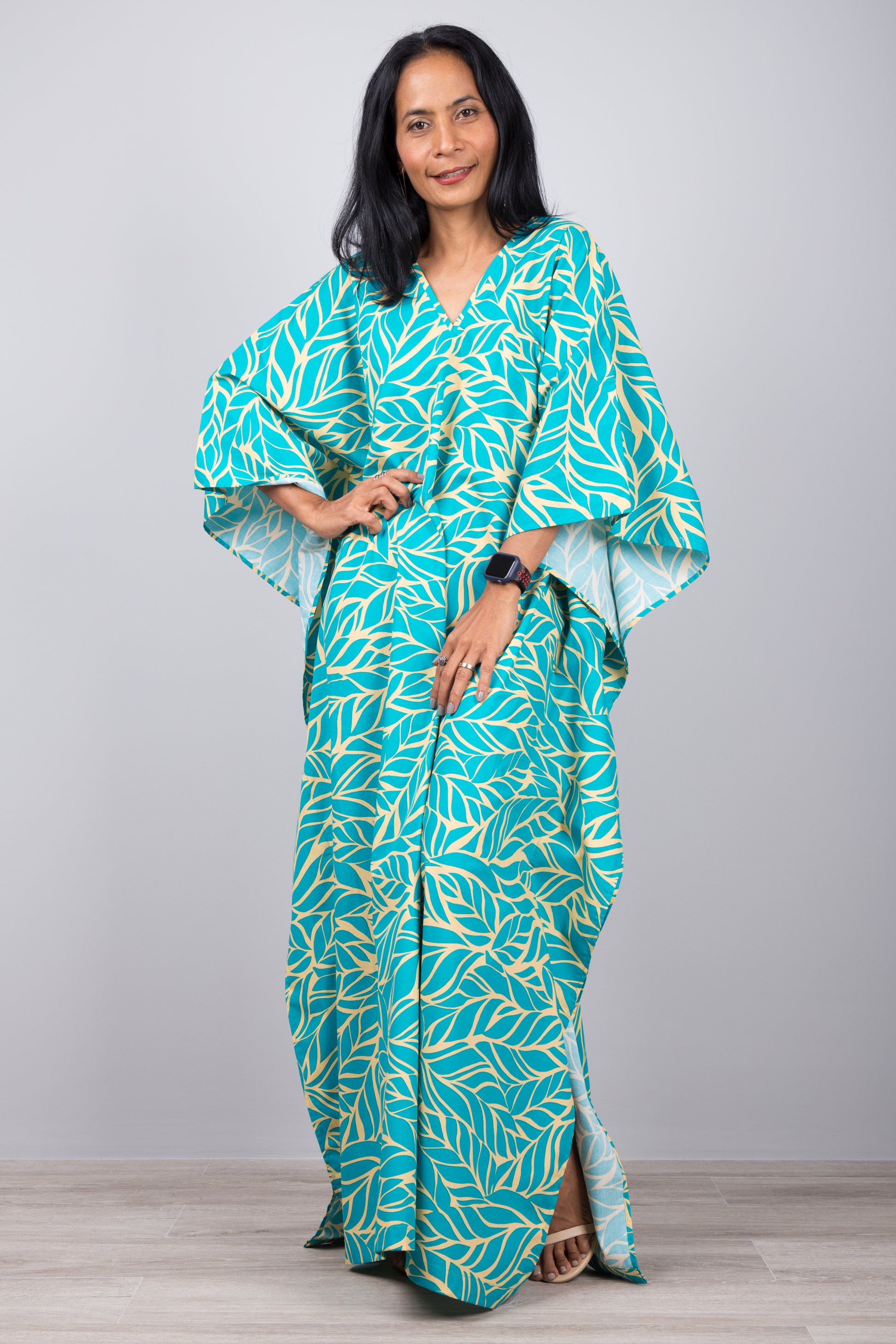 Woman in a vibrant turquoise kaftan dress with yellow leaf print, posed against a gray wall with a wooden floor, wearing a smartwatch and red bracelet.

