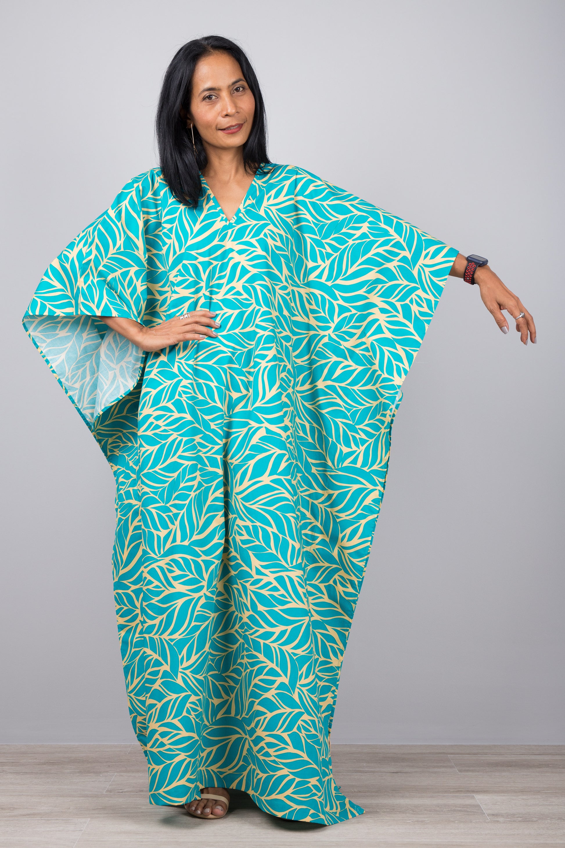 Woman in a tropical turquoise kaftan dress with yellow turquoise design, posed against a gray wall with a wooden floor, wearing a smartwatch and delicate necklace.

