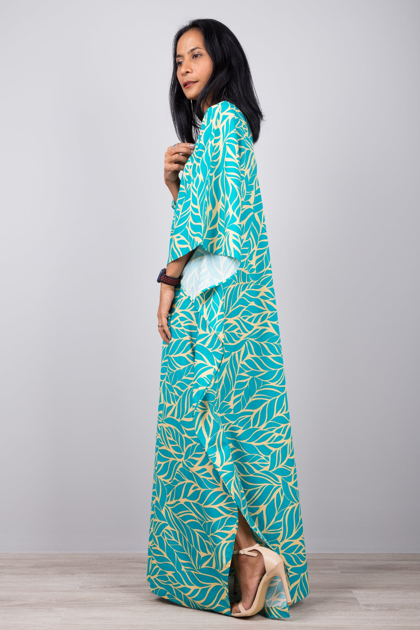 Woman in a boho-chic turquoise kaftan dress with tropical yellow leaf design, standing against a gray wall with a wooden floor, accessorized with a smartwatch and earrings

