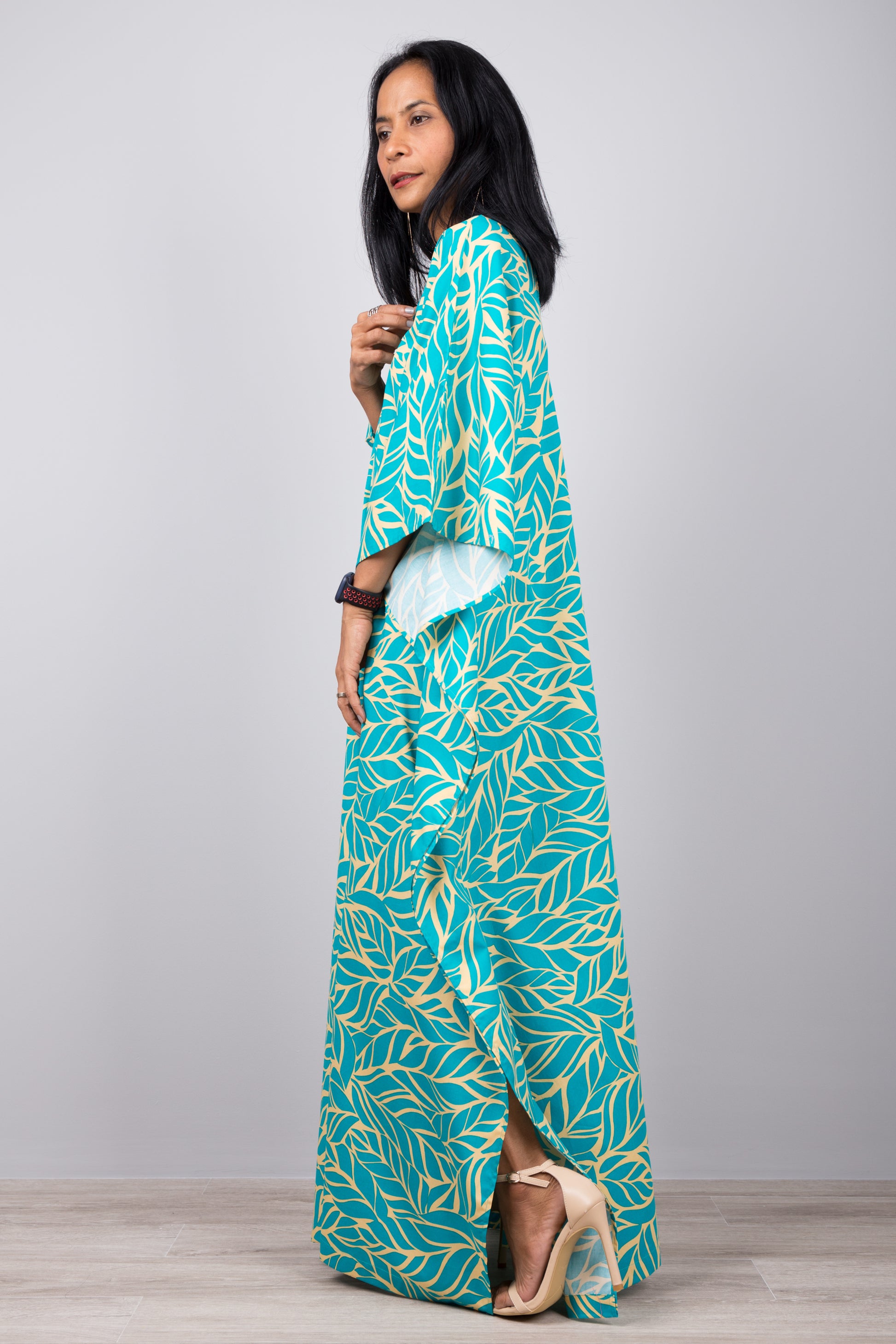 Woman in a boho-chic turquoise kaftan dress with tropical yellow leaf design, standing against a gray wall with a wooden floor, accessorized with a smartwatch and earrings

