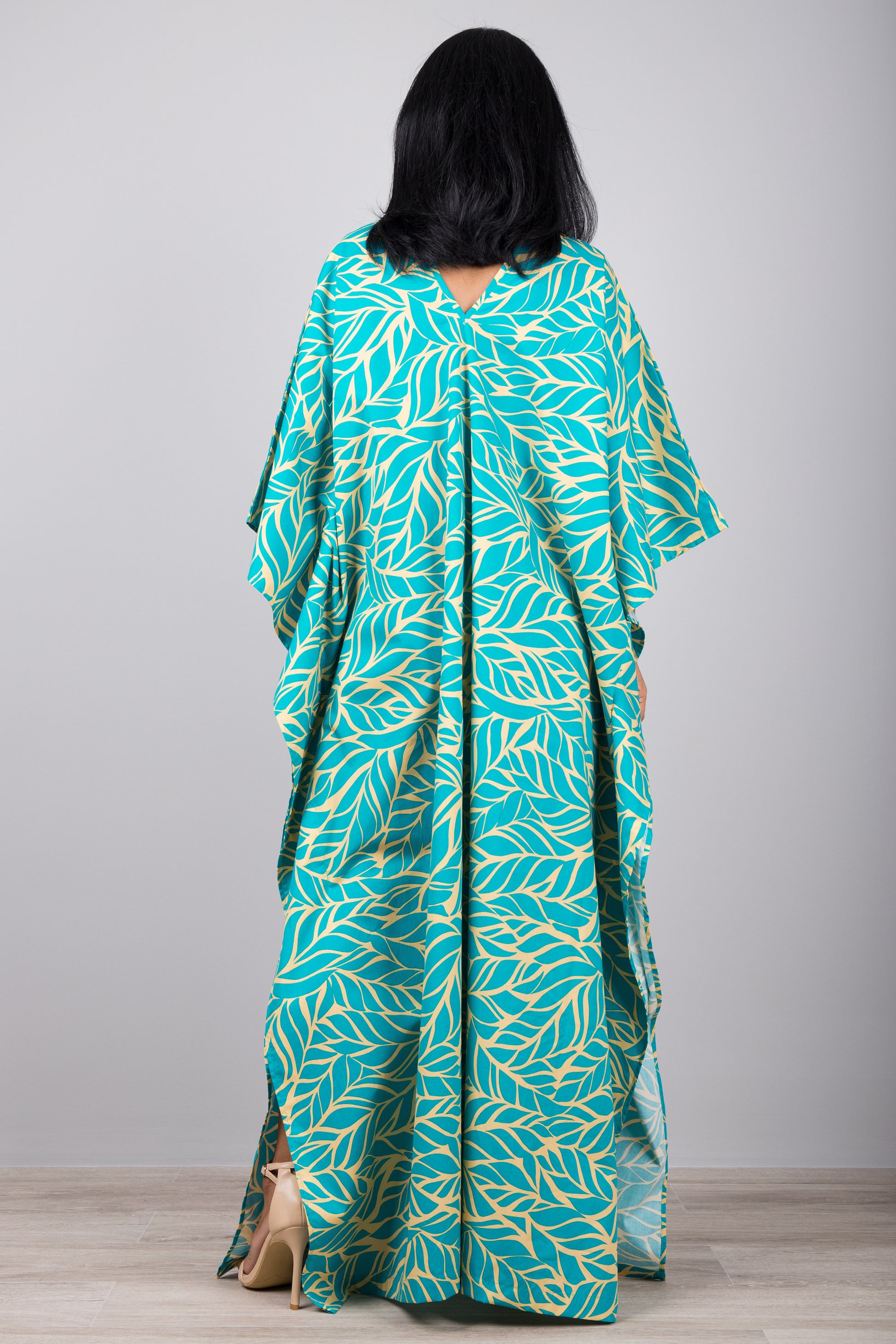 Woman in a lightweight turquoise kaftan dress with yellow leaf pattern, set against a gray wall with a wooden floor, wearing a smartwatch and silver cuff.

