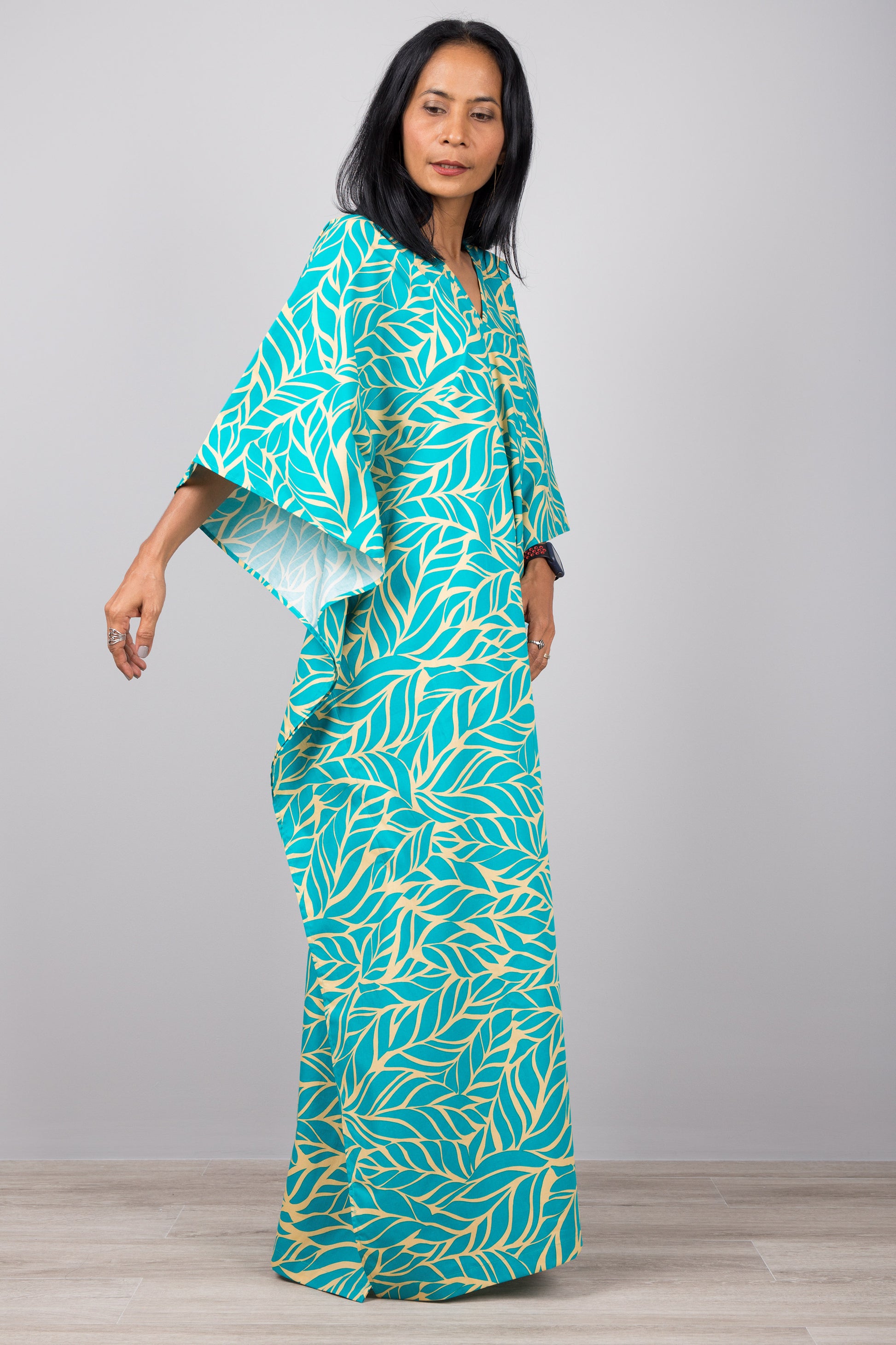 Woman in a flowy turquoise kaftan dress with yellow leaf print, standing against a gray wall with a wooden floor, accessorized with a smartwatch and minimalist jewelry.

