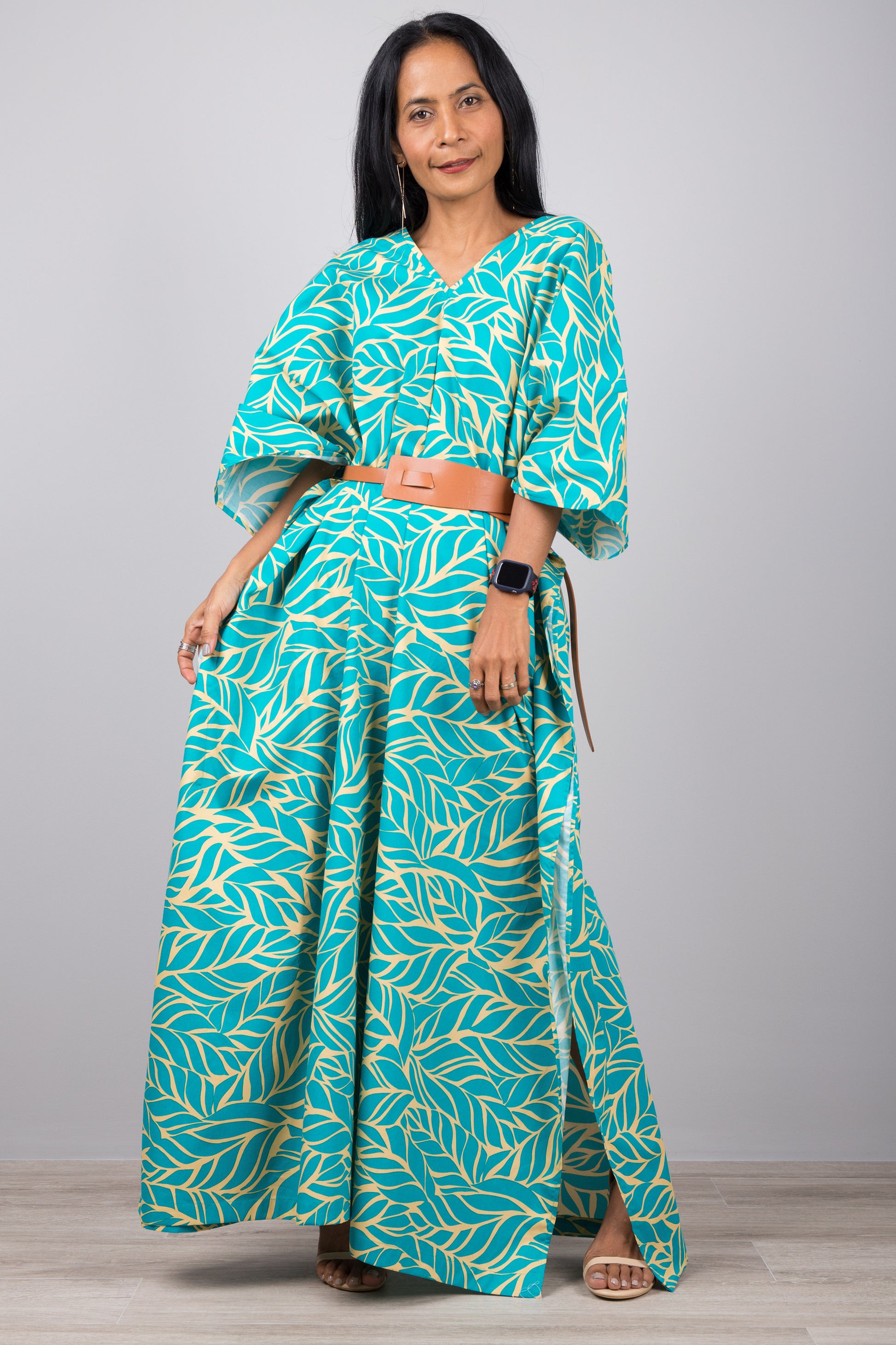Woman in a one-size turquoise kaftan dress with tropical yellow leaf pattern, posed against a gray wall with a wooden floor, wearing a smartwatch and strappy sandals


