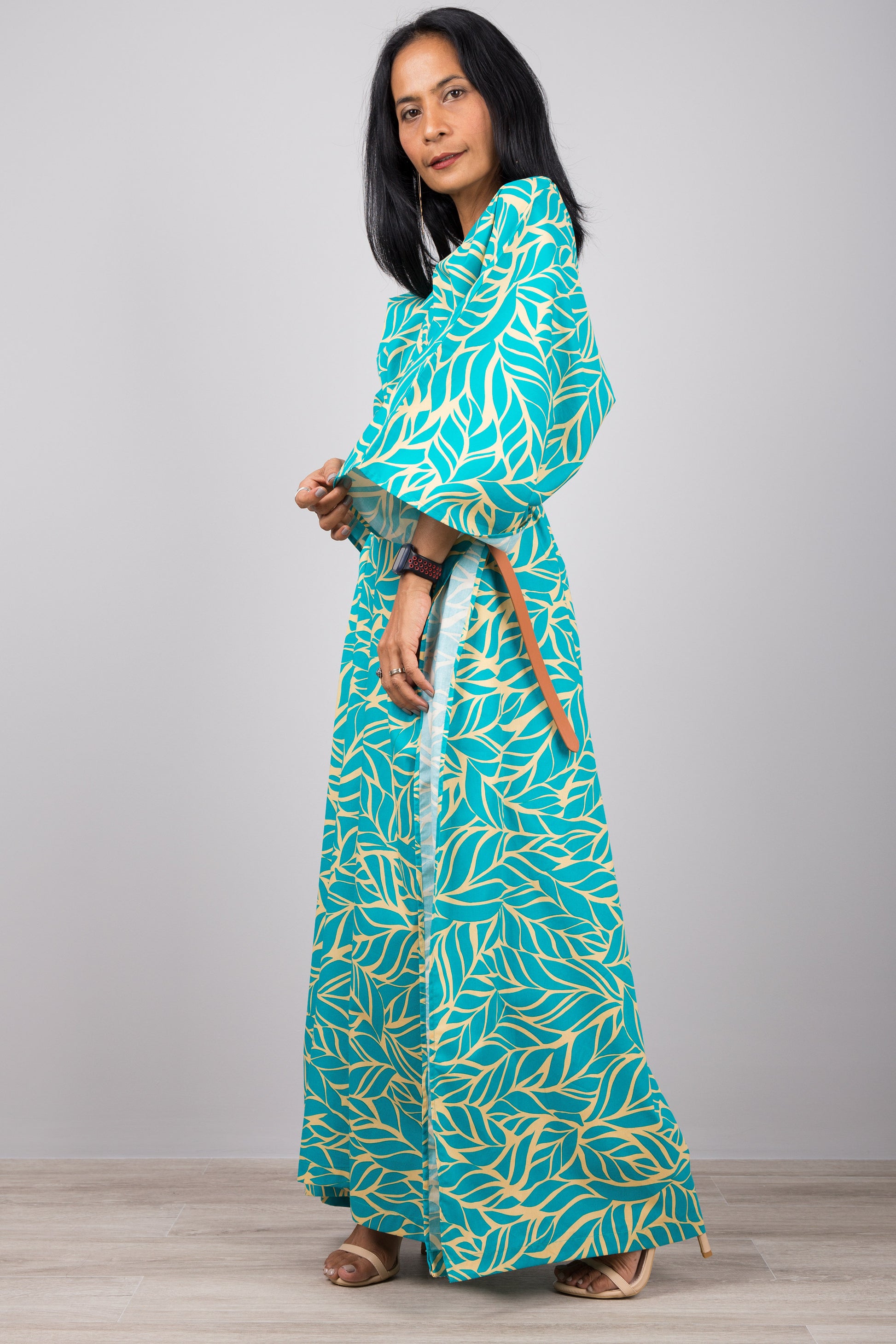 Woman in an elegant turquoise kaftan dress with yellow leaf design, standing against a gray wall with a wooden floor, accessorized with a smartwatch and ring

