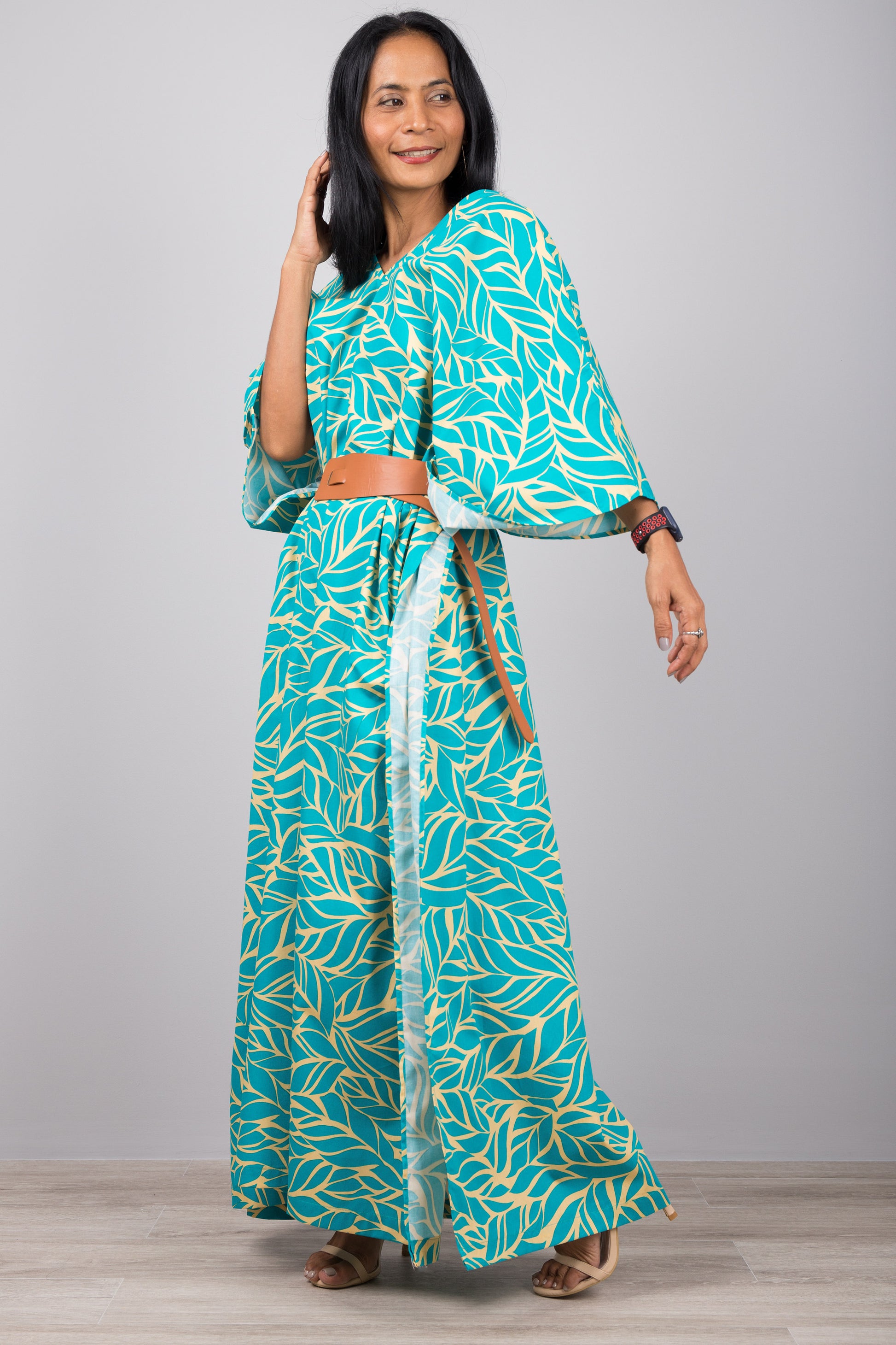 Woman in a summer-ready turquoise kaftan dress with yellow leaf print, set against a gray wall with a wooden floor, wearing a smartwatch and silver earrings

