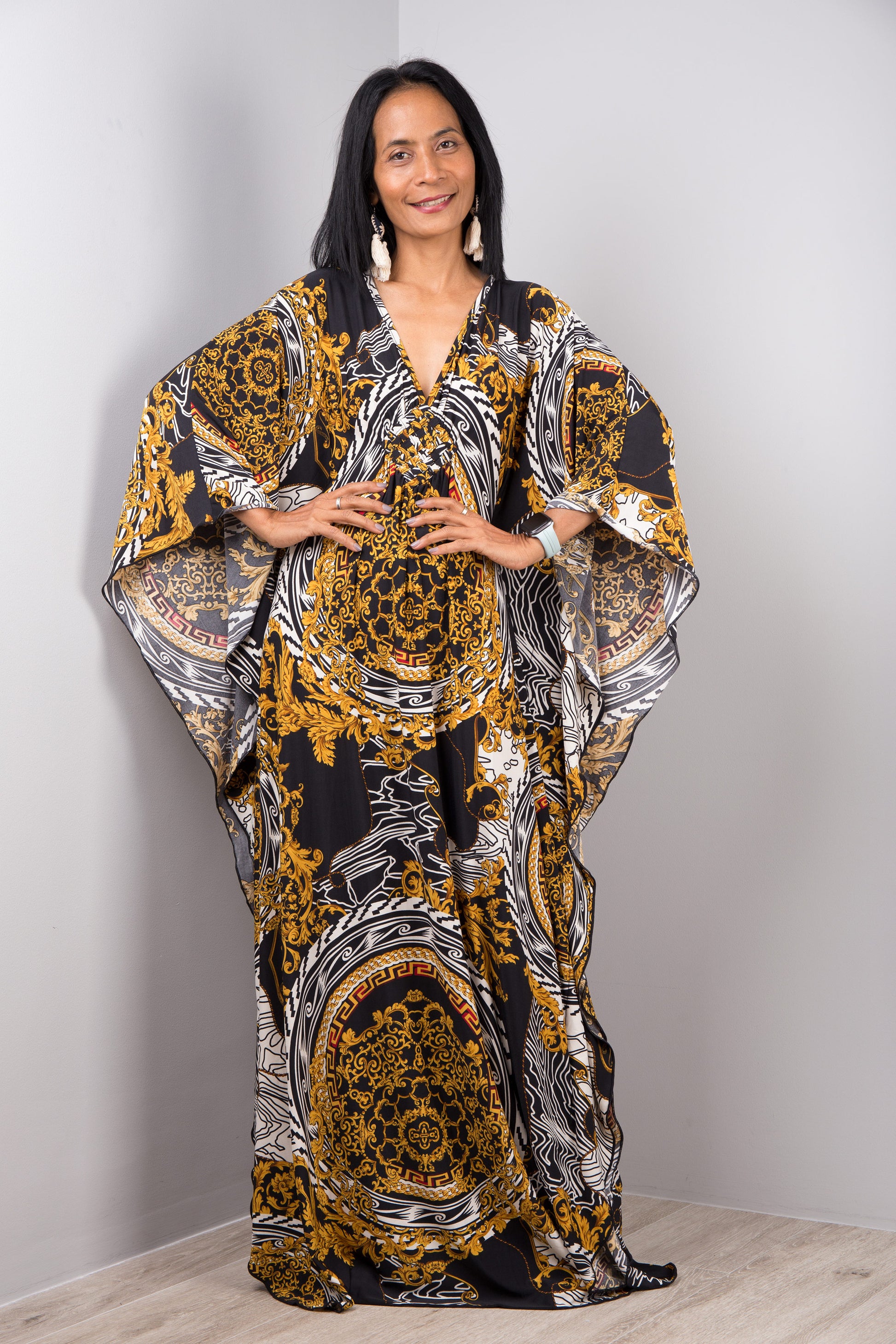 Woman in a black, gold, and white baroque-patterned kaftan dress by Nuichan, standing with hands on hips, showcasing the dress’s wide sleeves and maxi length, in a studio with a gray backdrop and wooden floor.