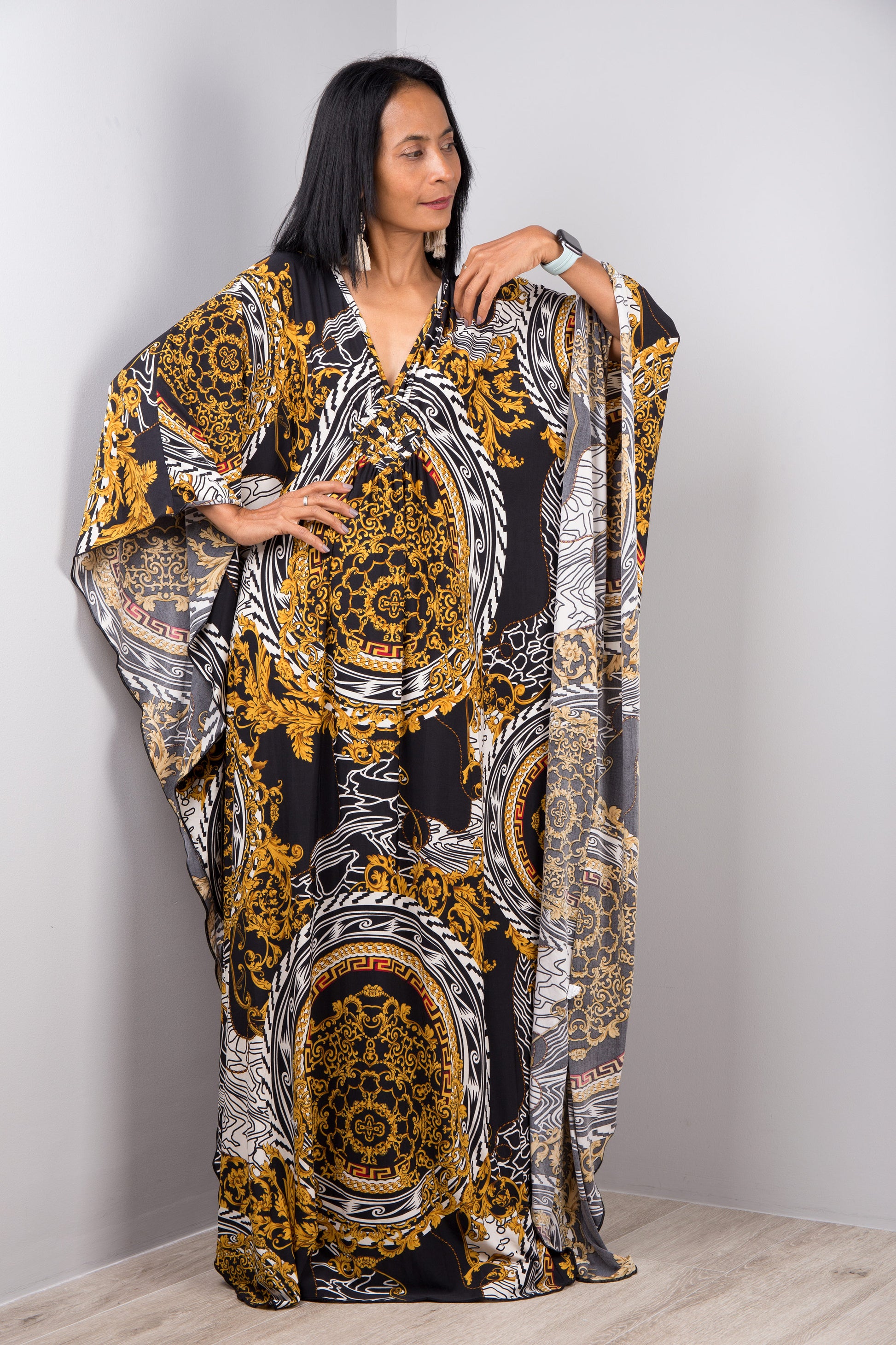 Nuichan’s black and gold baroque kaftan dress with intricate white accents, modeled by a woman with long black hair, posed gracefully in a minimalist studio with a gray background and light wood flooring.