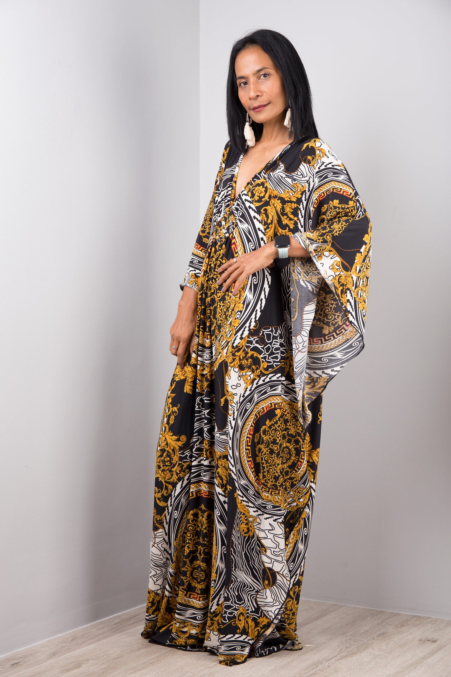 A woman wearing Nuichan’s black and gold baroque kaftan dress with white accents, standing sideways in a minimalist studio, showcasing the dress’s loose fit, wide sleeves, and maxi length against a gray wall and wooden floor.