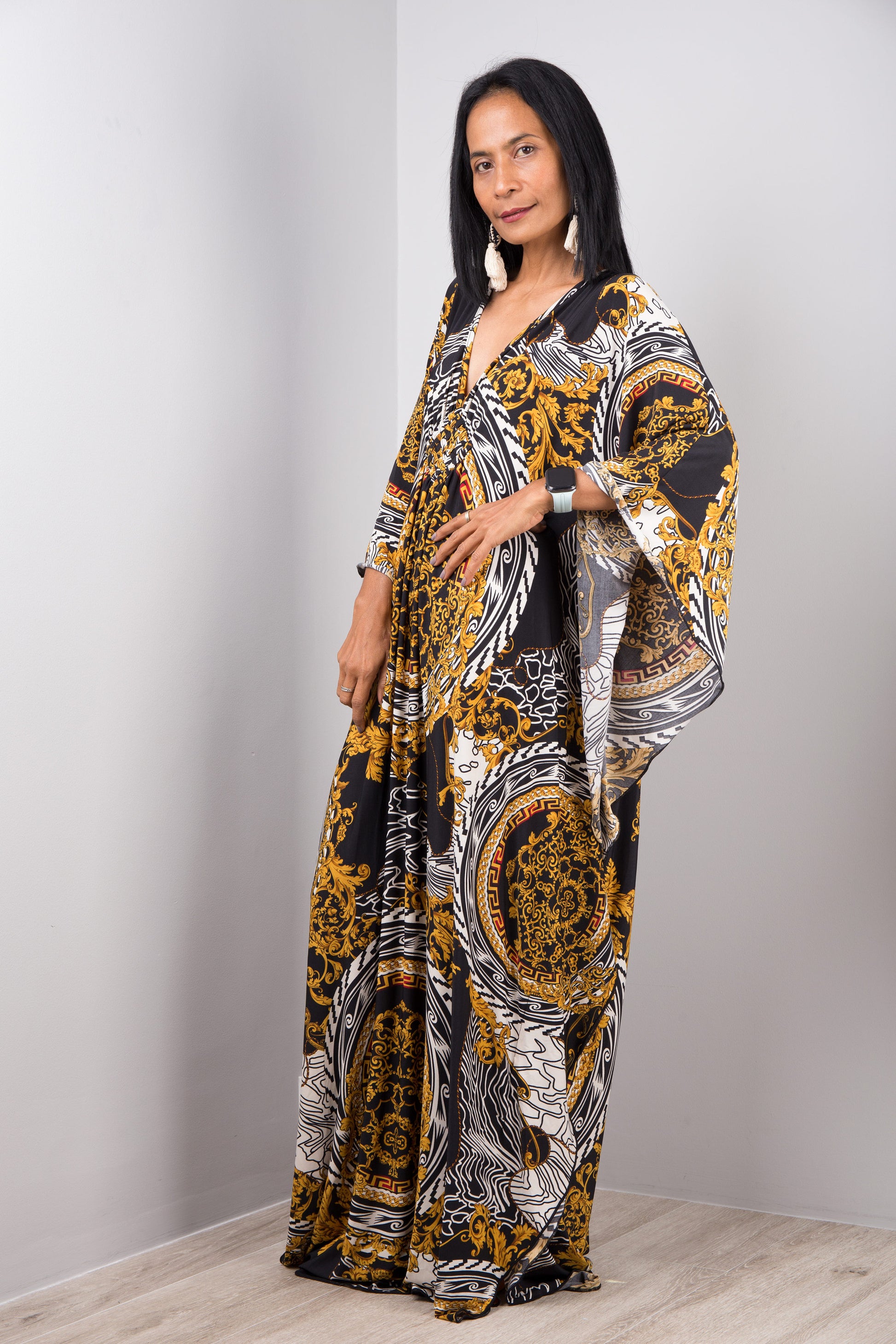 A woman wearing Nuichan’s black and gold baroque kaftan dress with white accents, standing sideways in a minimalist studio, showcasing the dress’s loose fit, wide sleeves, and maxi length against a gray wall and wooden floor.
