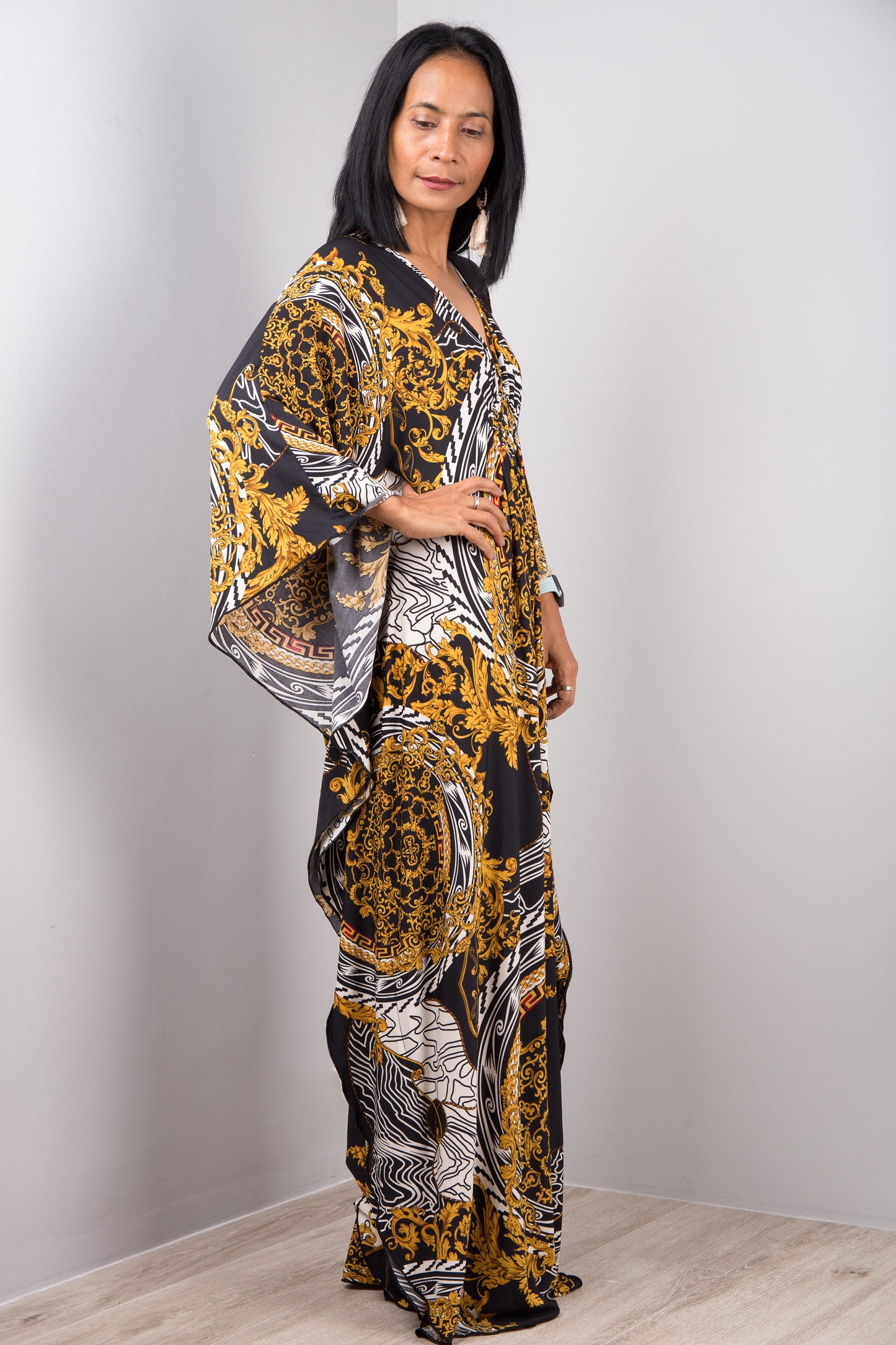 A woman wearing Nuichan’s black and gold baroque kaftan dress with white accents, standing sideways in a minimalist studio, showcasing the dress’s loose fit, wide sleeves, and maxi length against a light gray wall and wooden floor.