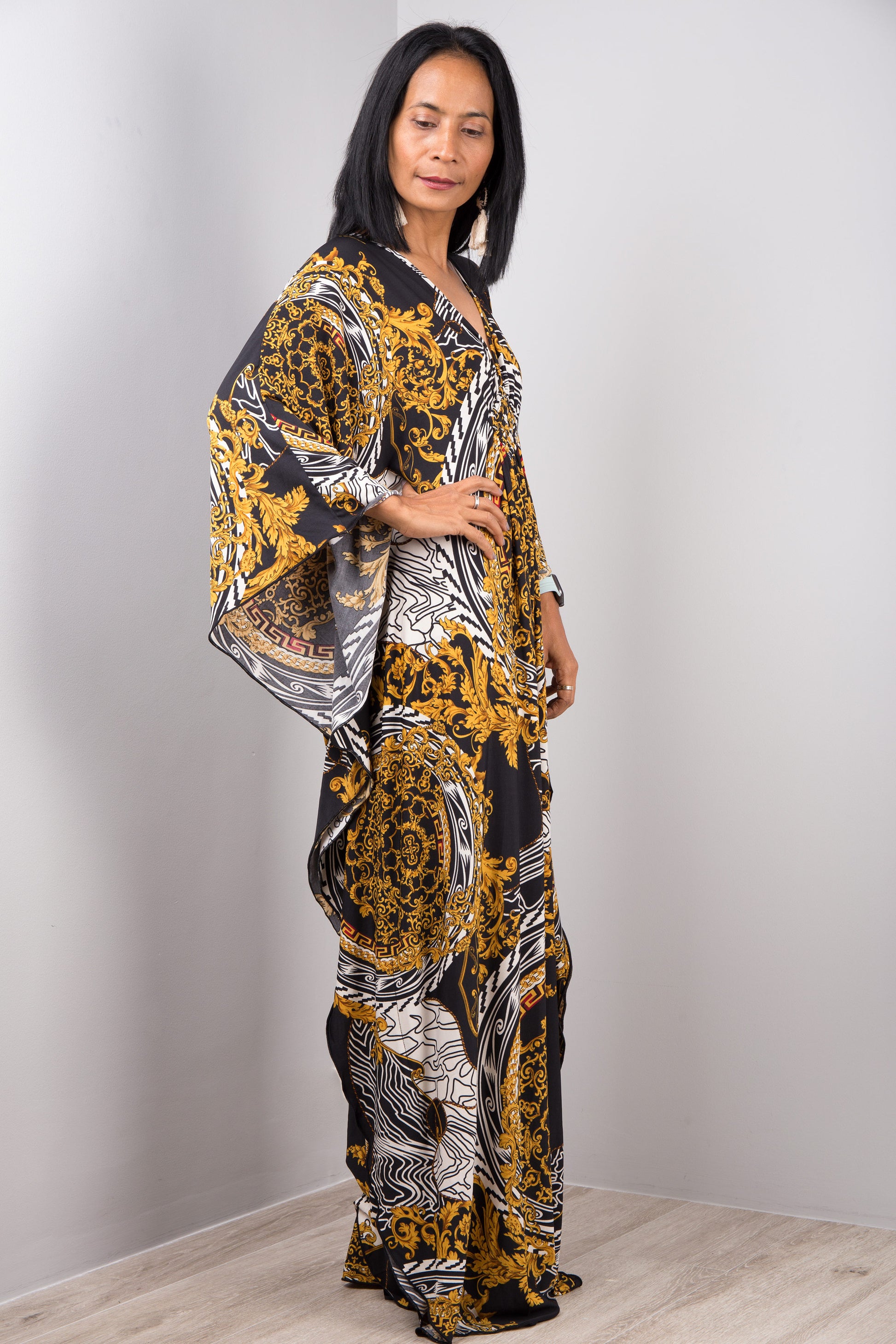 A woman wearing Nuichan’s black and gold baroque kaftan dress with white accents, standing sideways in a minimalist studio, showcasing the dress’s loose fit, wide sleeves, and maxi length against a light gray wall and wooden floor.