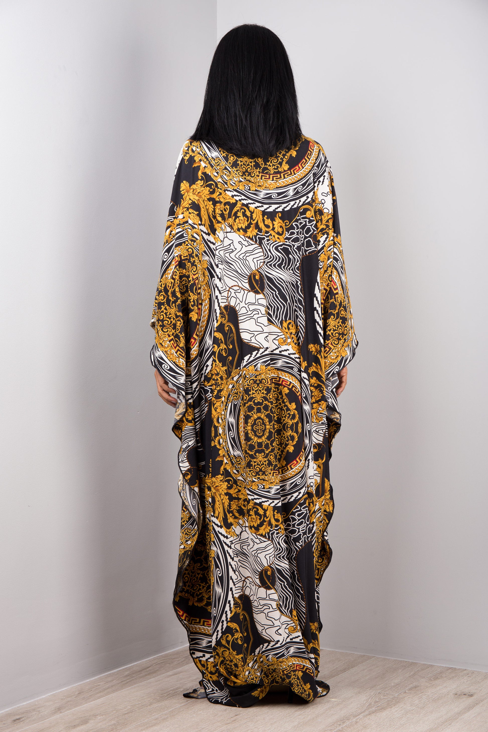 A woman wearing Nuichan’s black and gold baroque kaftan dress with white accents, backside view in a minimalist studio, showcasing the dress’s loose fit, wide sleeves, and maxi length against a gray wall and wooden floor.