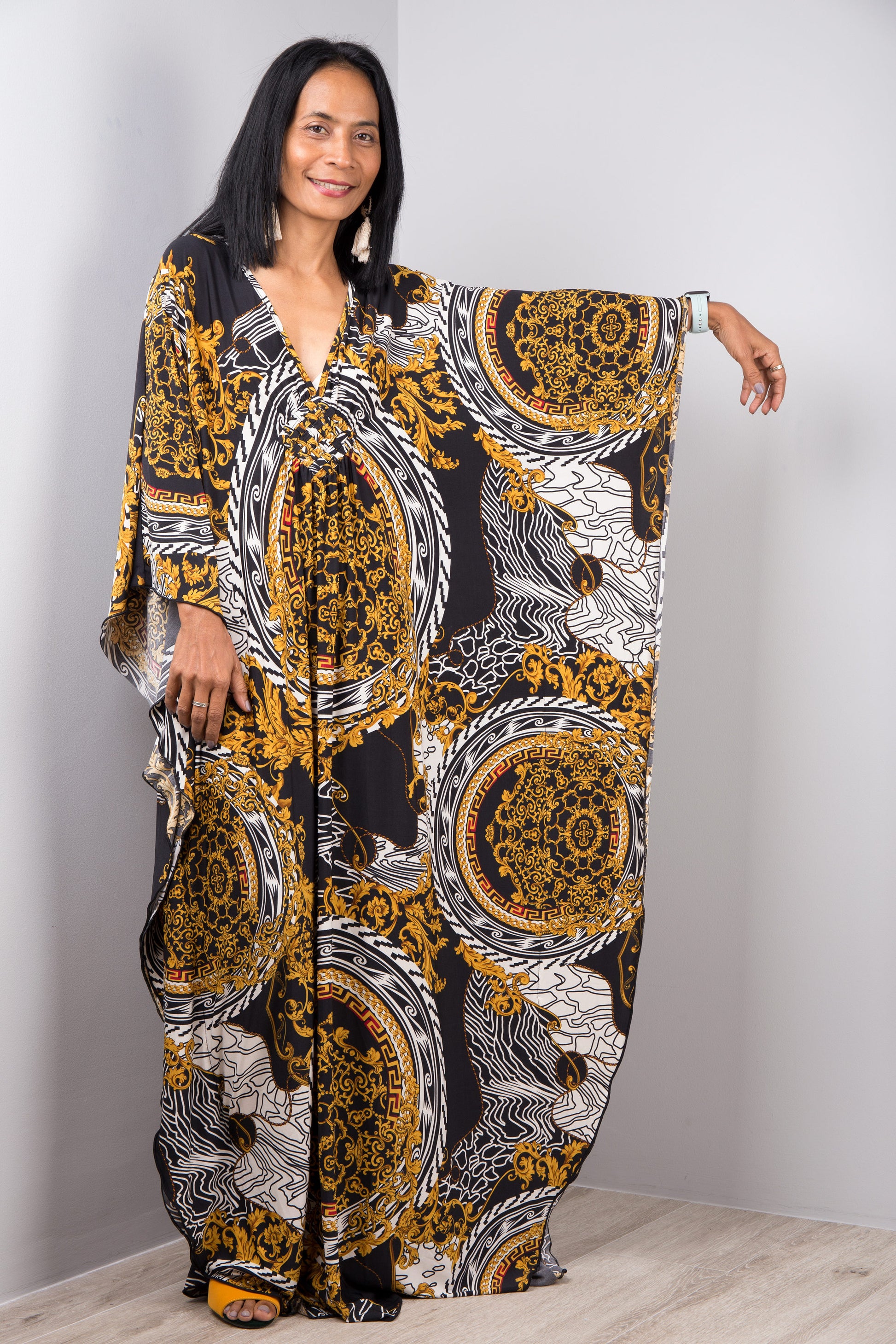 Black, gold, and white baroque-patterned kaftan dress from Nuichan, worn by a woman with dark hair and a white smartwatch, posed against a gray studio wall and light wood floor, highlighting its opulent boho-chic design and flowing silhouette.