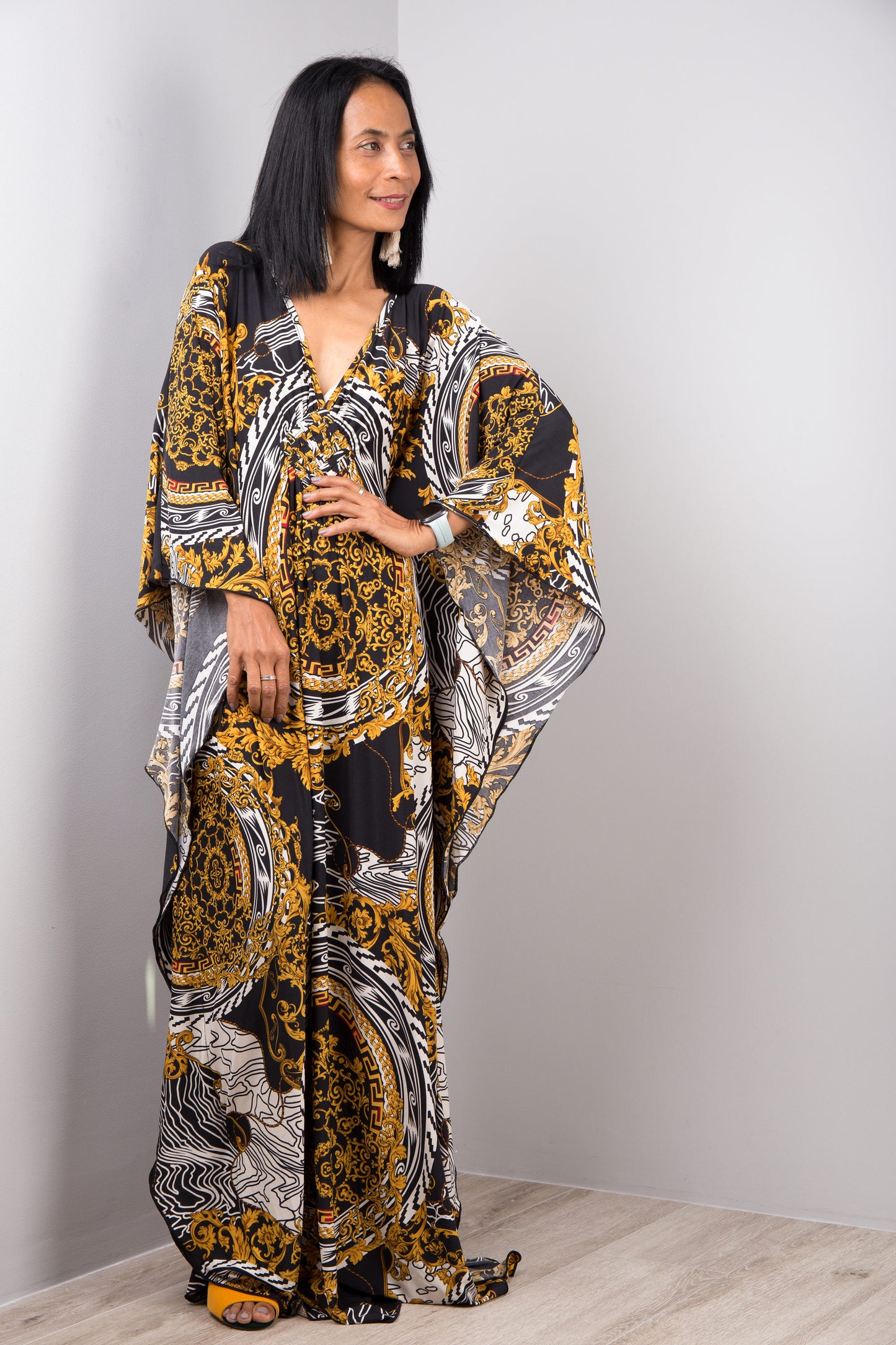 A woman modeling a black, gold, and white baroque-patterned kaftan dress from Nuichan, with a loose fit, V-neck, and flowing sleeves, standing in a simple gray and wood studio setting, accessorized with earrings.