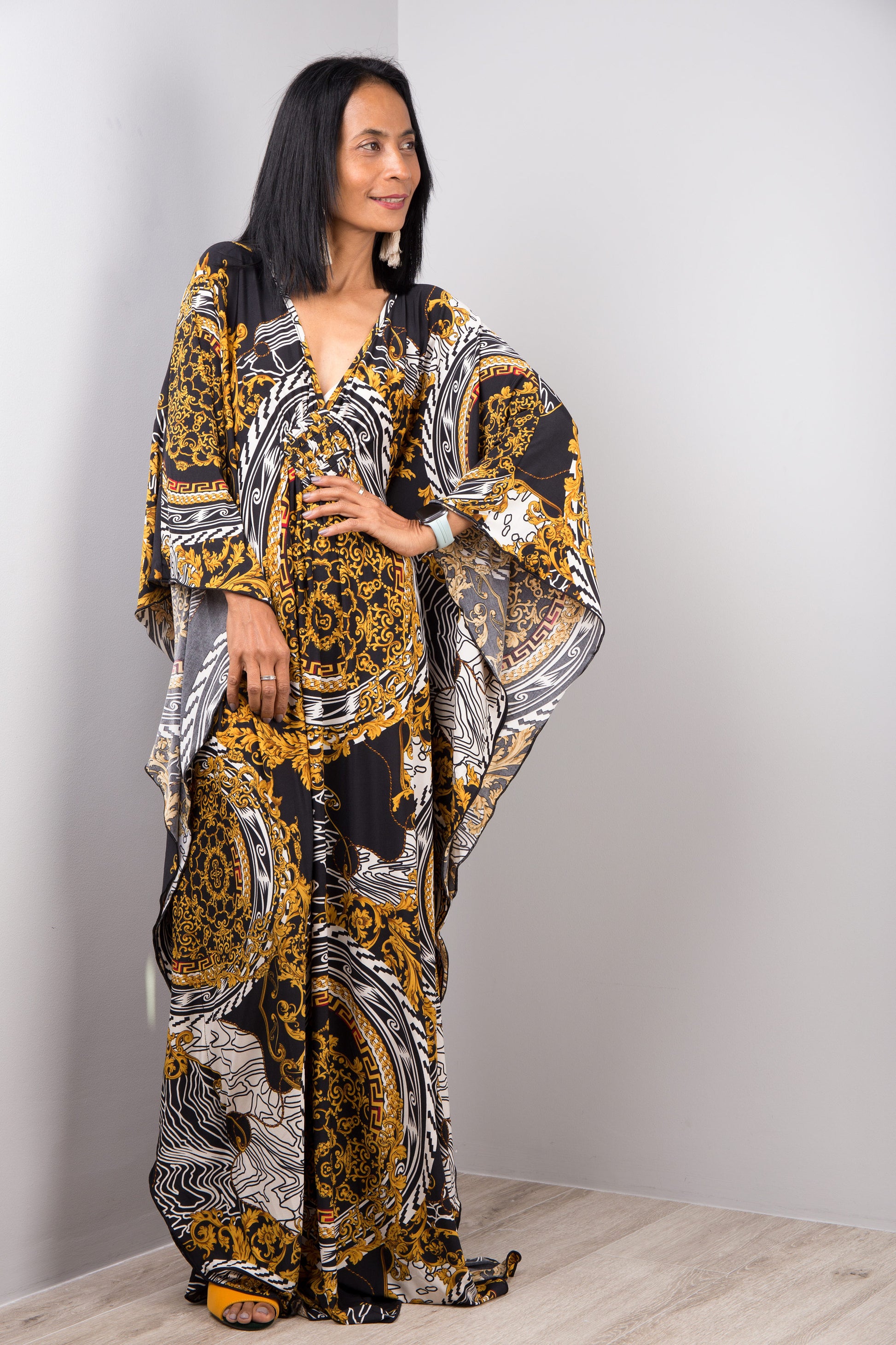A woman modeling a black, gold, and white baroque-patterned kaftan dress from Nuichan, with a loose fit, V-neck, and flowing sleeves, standing in a simple gray and wood studio setting, accessorized with earrings.