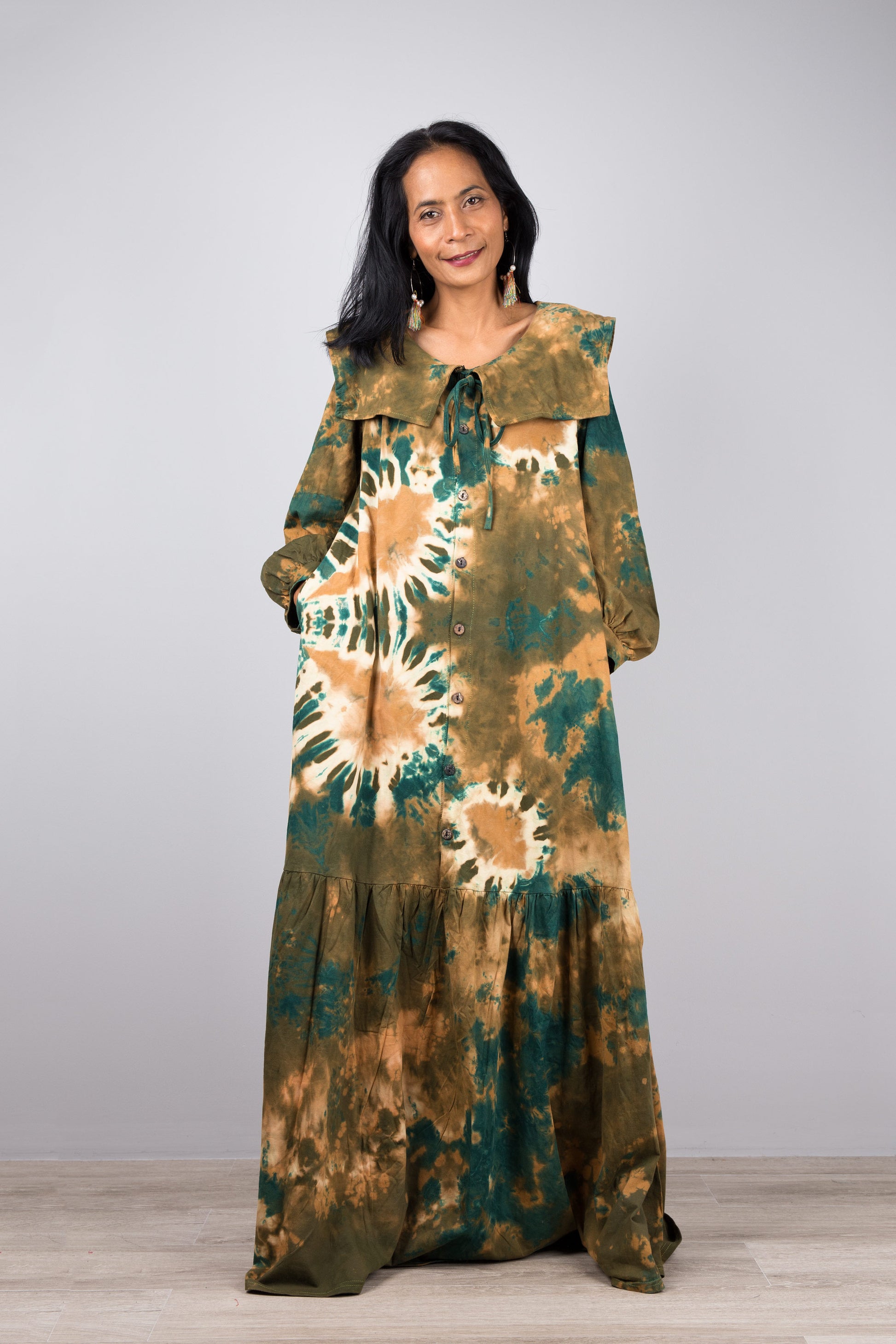 Stylish woman in a floor-length green tie-dye kaftan dress with navy trim, V-neck, and billowy sleeves, posed against a gray wall and wooden floor, wearing nude sandals and earrings


