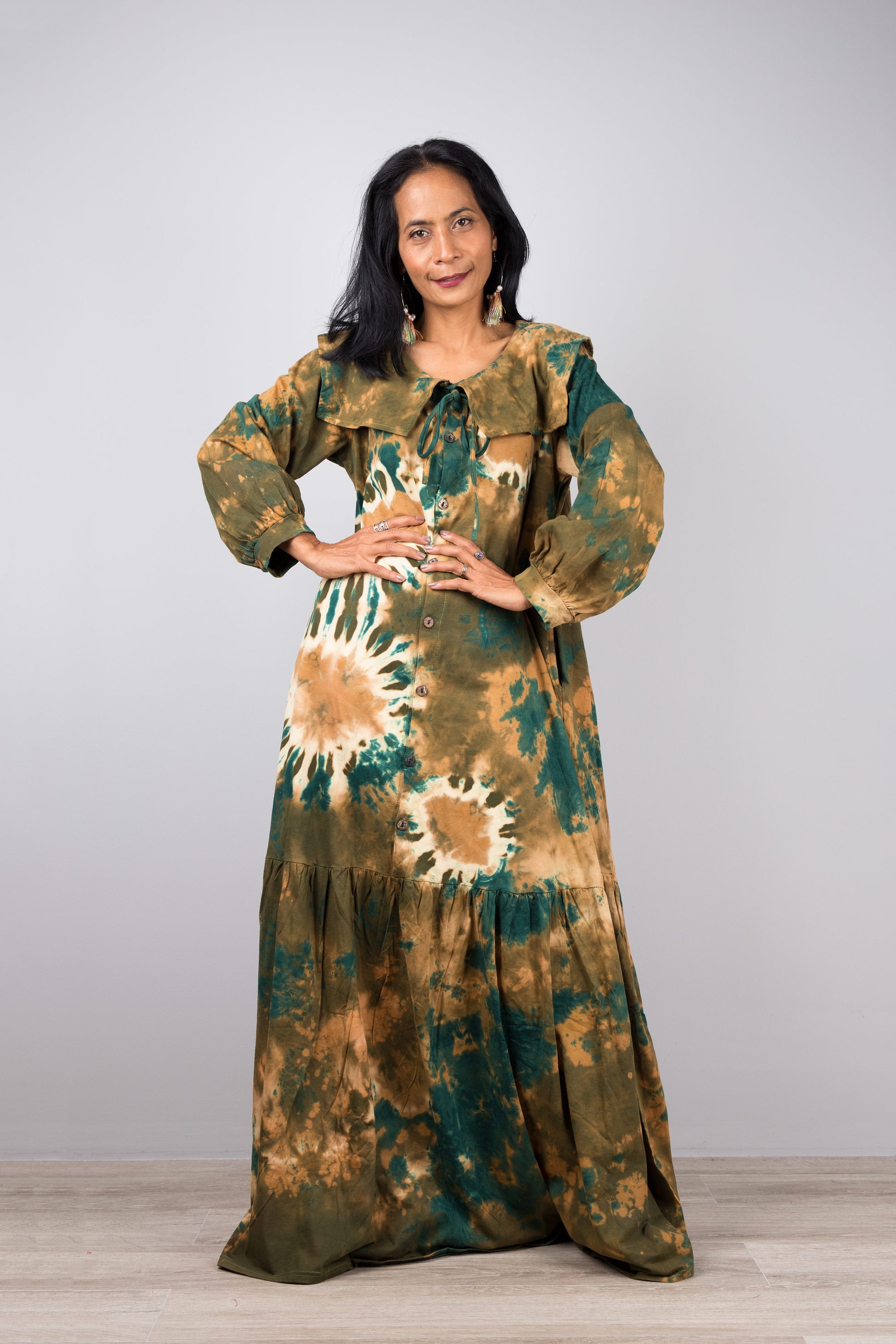 Stylish woman in a floor-length green tie-dye kaftan dress with navy trim, V-neck, and billowy sleeves, posed against a gray wall and wooden floor, wearing nude sandals and earrings

