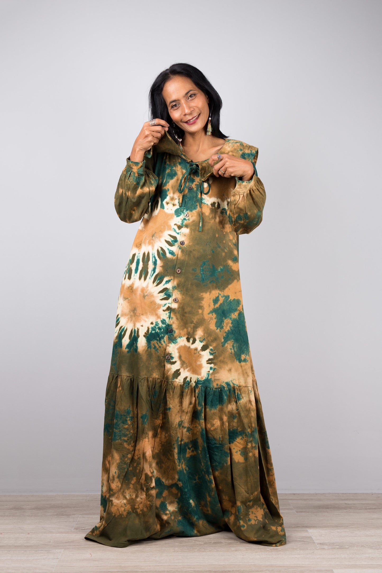 Woman showcasing a green tie-dye kaftan maxi dress with navy trim, flowing design, V-neck, and long sleeves, accessorized with nude sandals, on a gray and wooden floor setting

