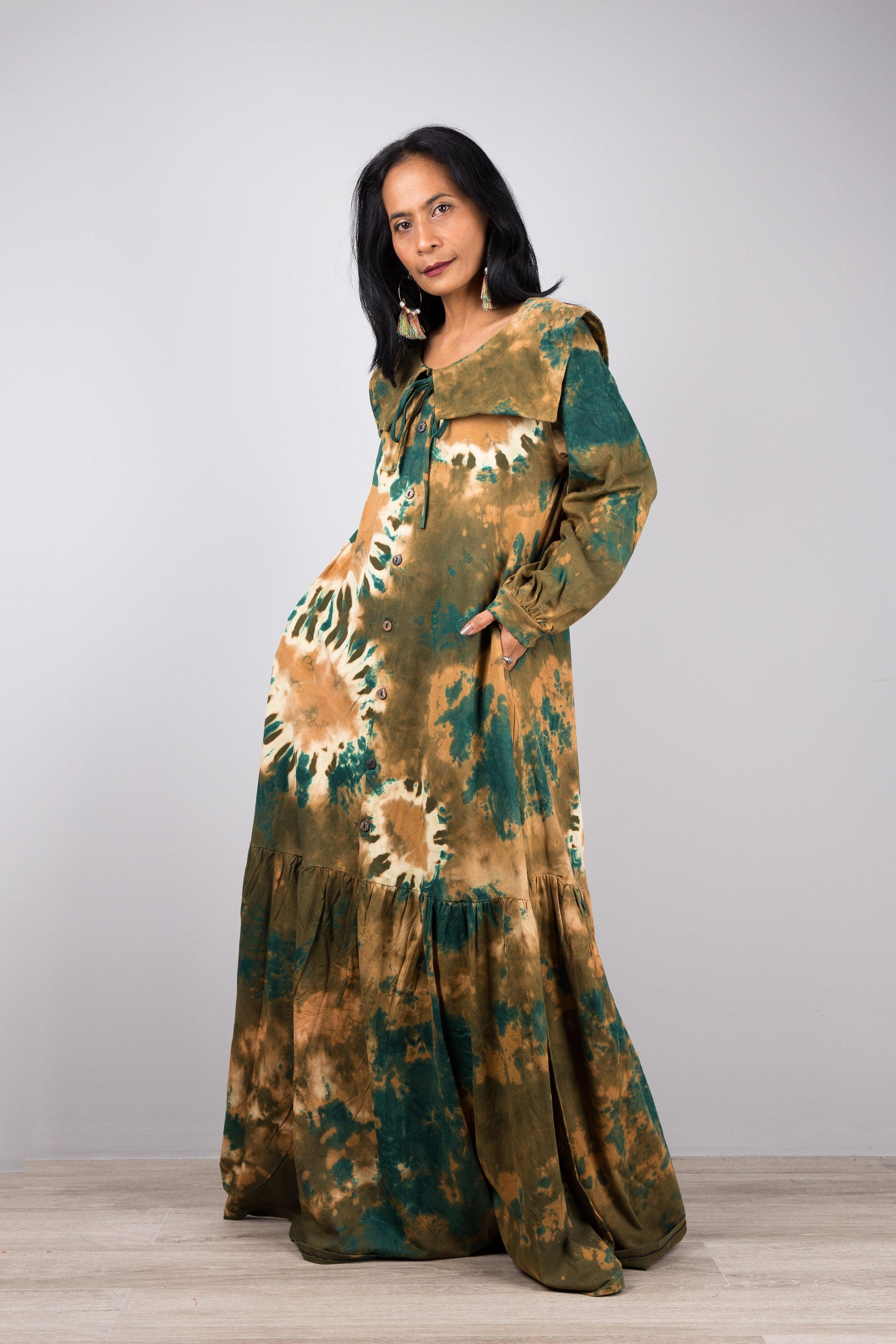 Elegant green tie-dye kaftan maxi dress with a navy-trimmed collar and gathered sleeves, worn by a woman against a gray wall and wooden floor, paired with nude sandals and earrings.

