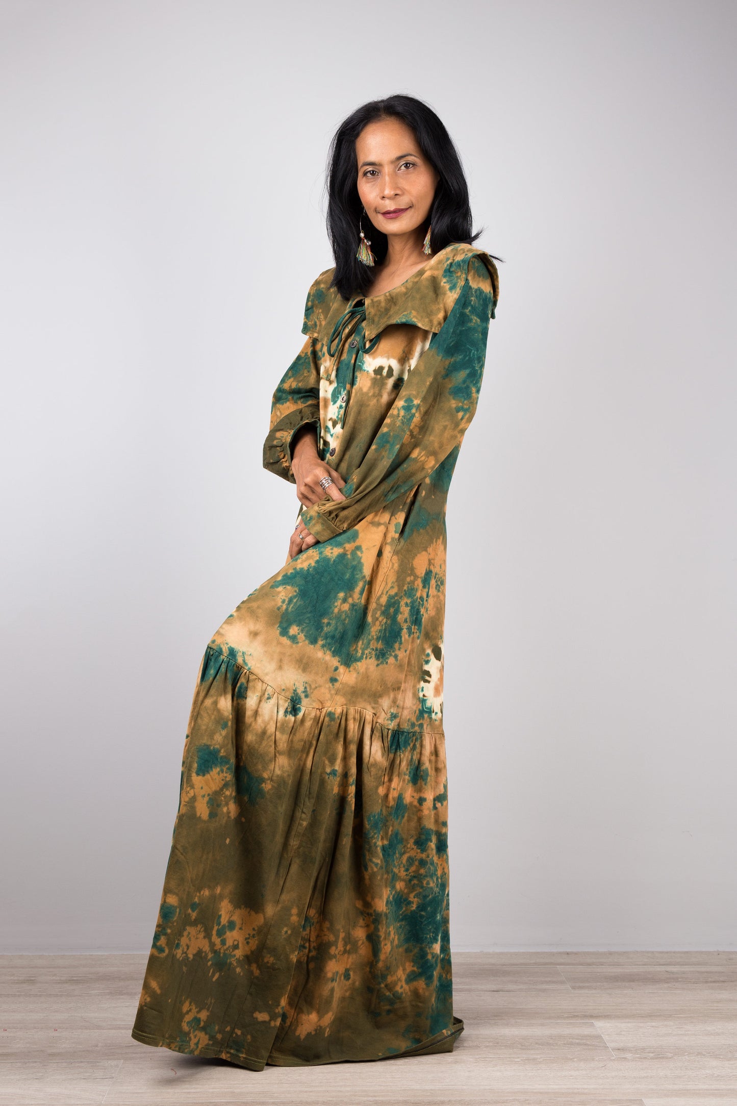Model in a green tie-dye pattern kaftan maxi dress with navy trim, deep V-neck, and long sleeves, standing on a wooden floor with a gray wall background, wearing nude sandals.

