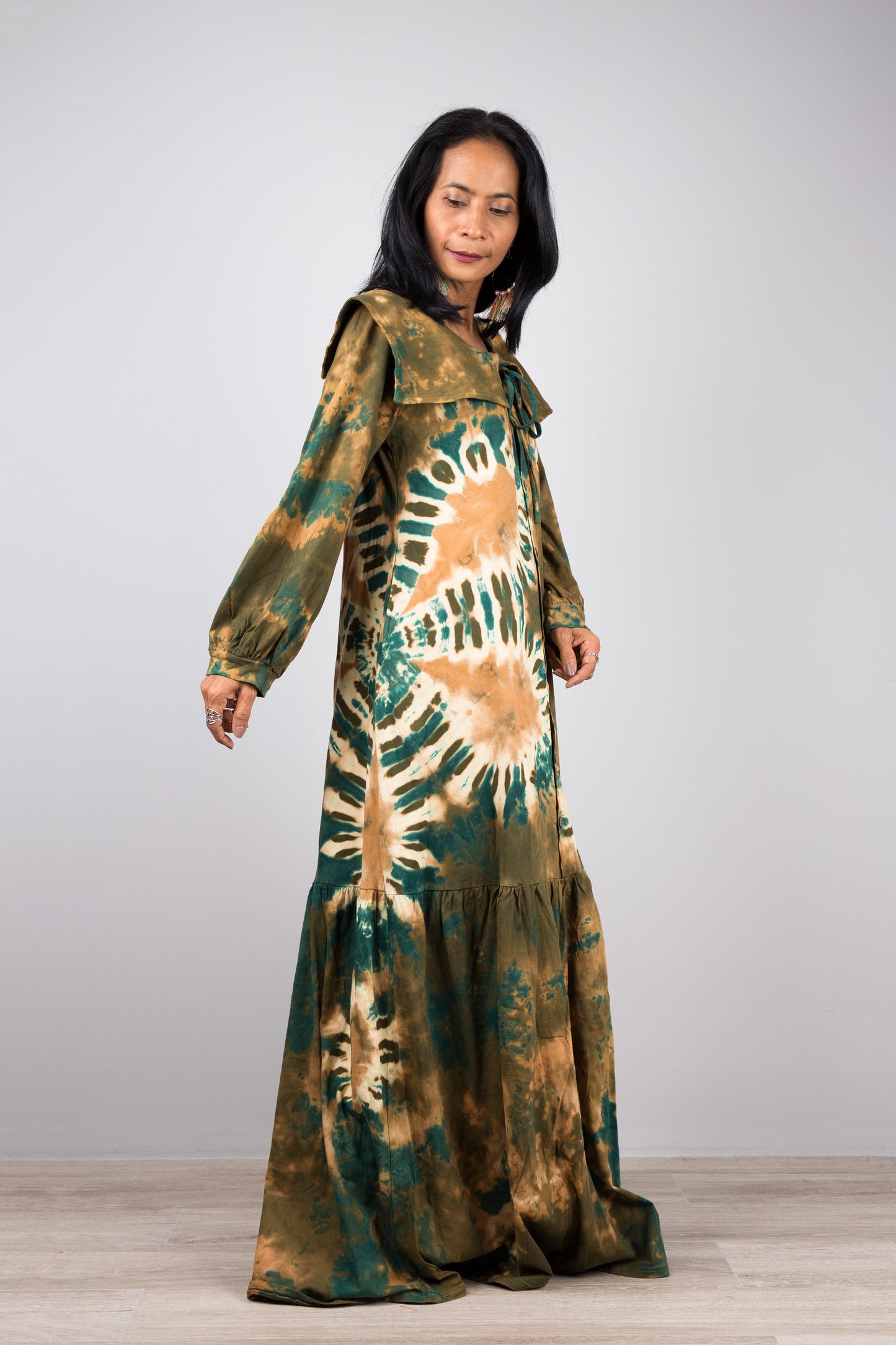 Woman in a vibrant green tie-dye kaftan maxi dress with navy collar and billowy sleeves, posed against a gray wall and wooden floor, styled with nude sandals and dangling earrings

