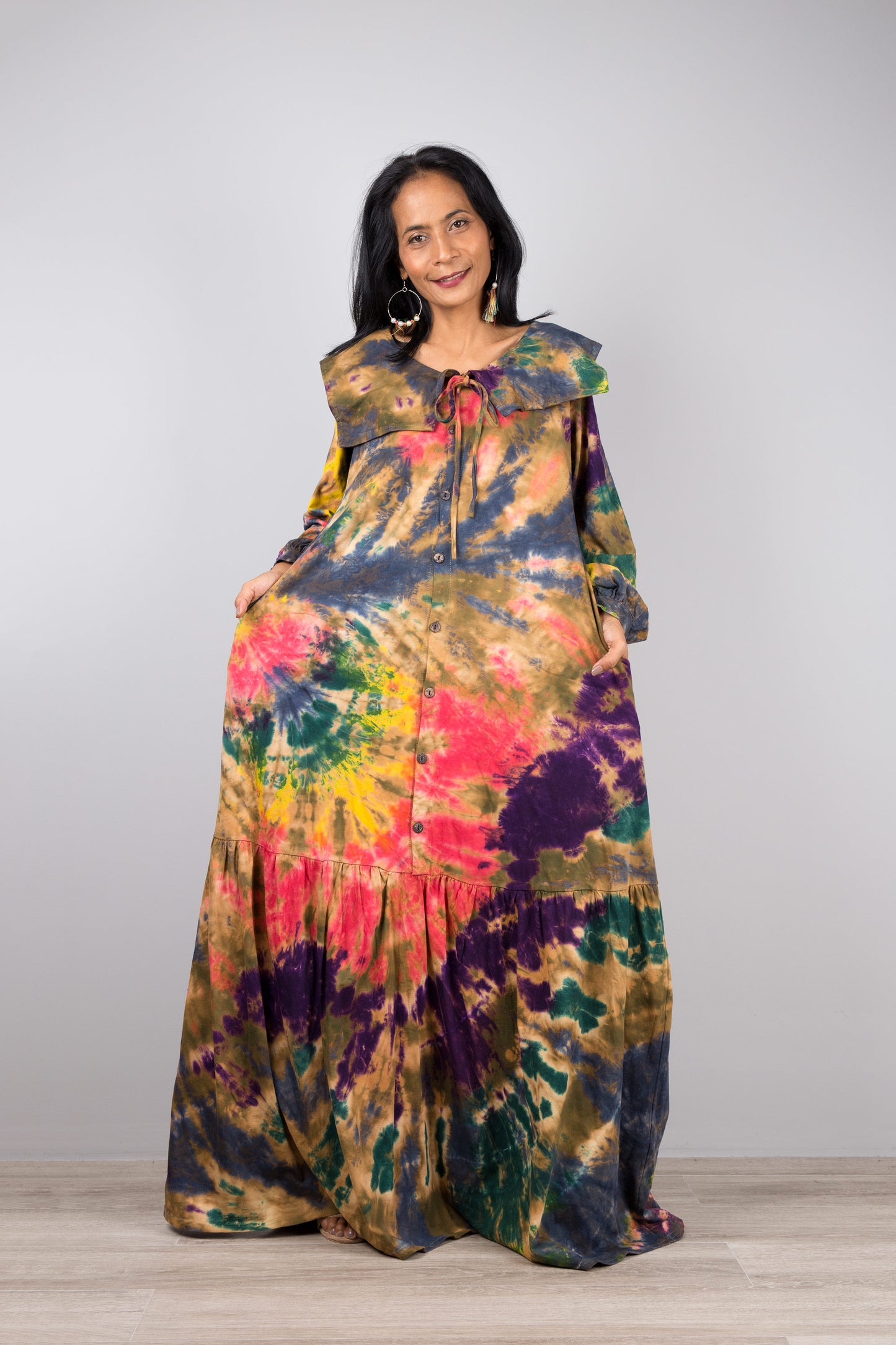 Woman showcasing a tie-dye kaftan maxi dress with navy trim, flowing design, deep V-neck, and long sleeves, accessorized with nude sandals, on a gray and wooden floor setting.


