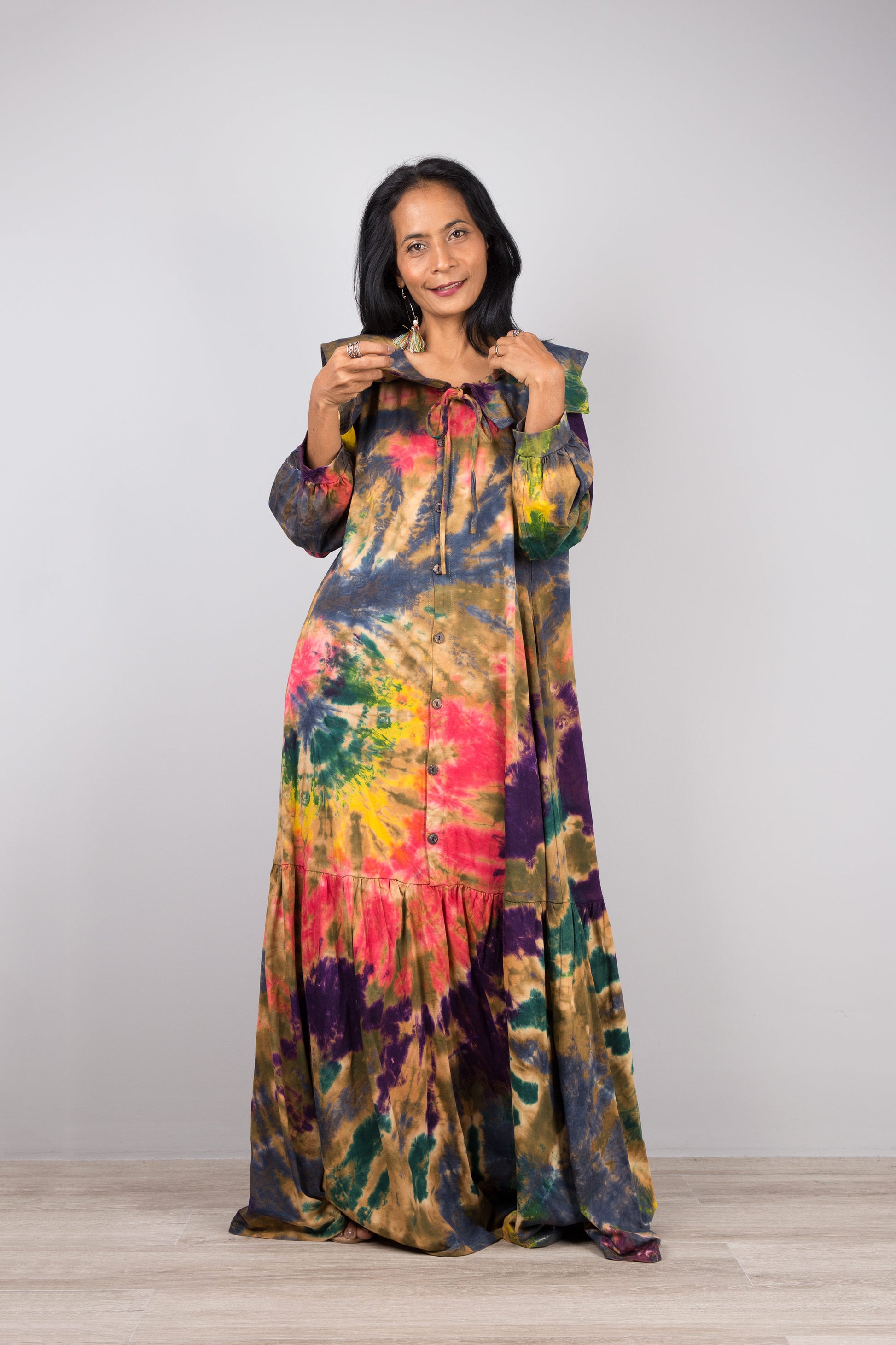 Nuichan tie-dye kaftan maxi dress with a navy-trimmed collar, deep V-neck, and long sleeves, modeled by a woman with nude sandals and dangling earrings, against a simple gray and wood backdrop.


