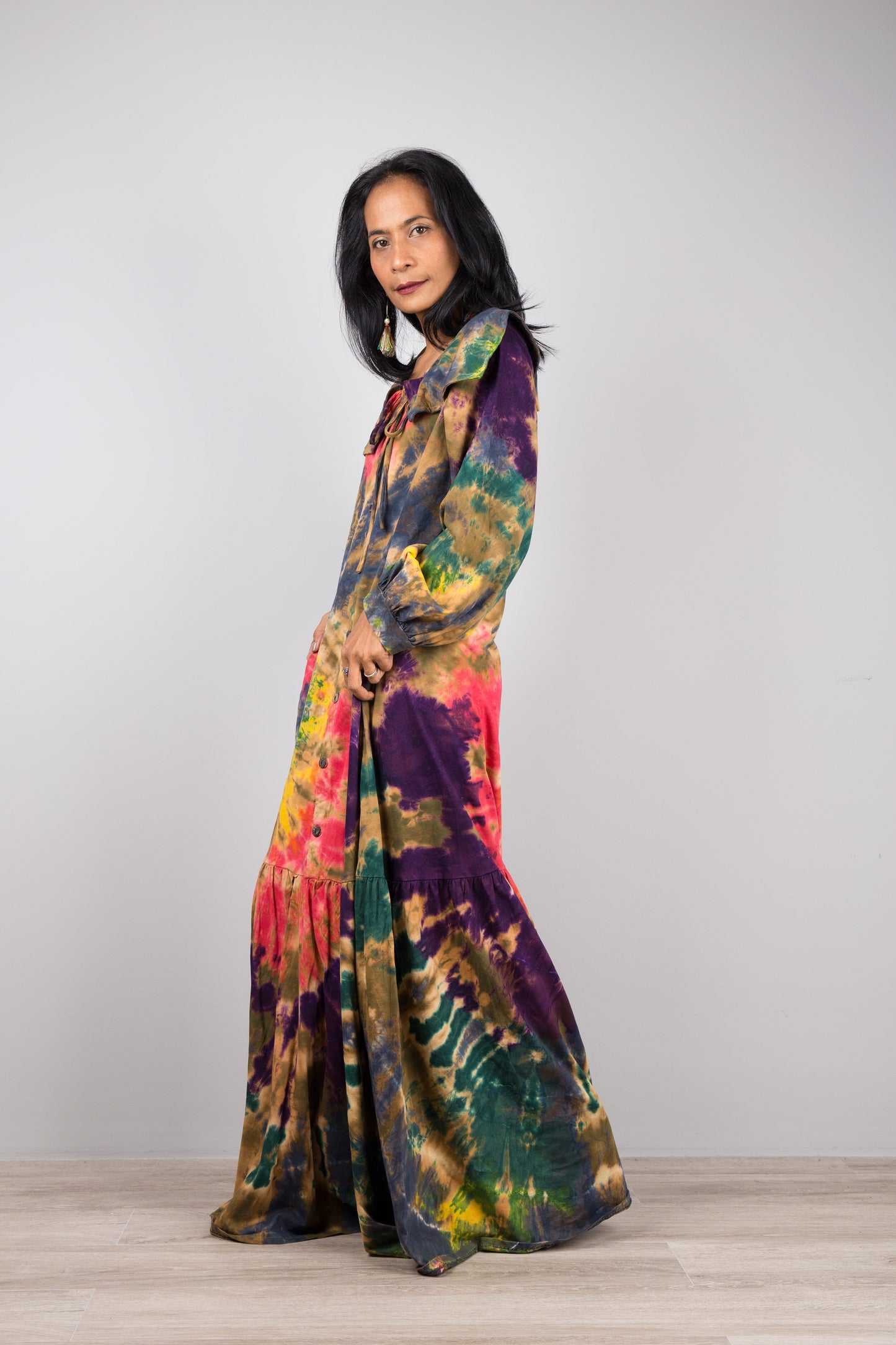 Model in a tie-dye pattern kaftan maxi dress with navy trim, deep V-neck, and long sleeves, standing on a wooden floor with a gray wall background, wearing nude sandals.

